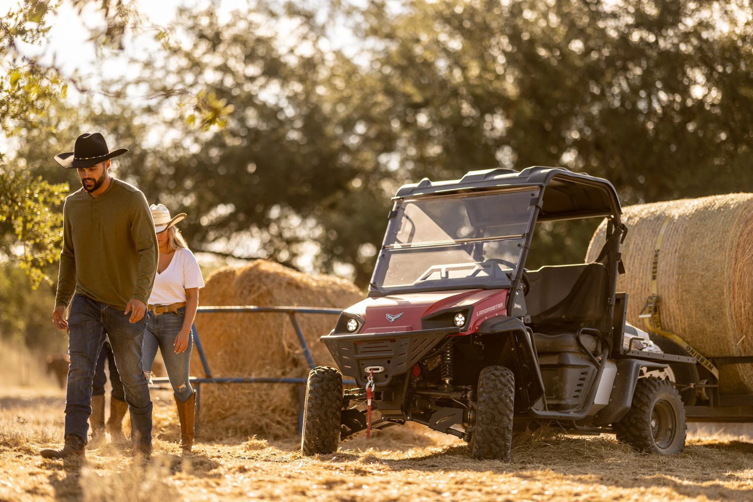 Two people, a man and a woman, walking outdoors during sunset, with hay bales and a utility vehicle in the background, in a rural or farm setting.