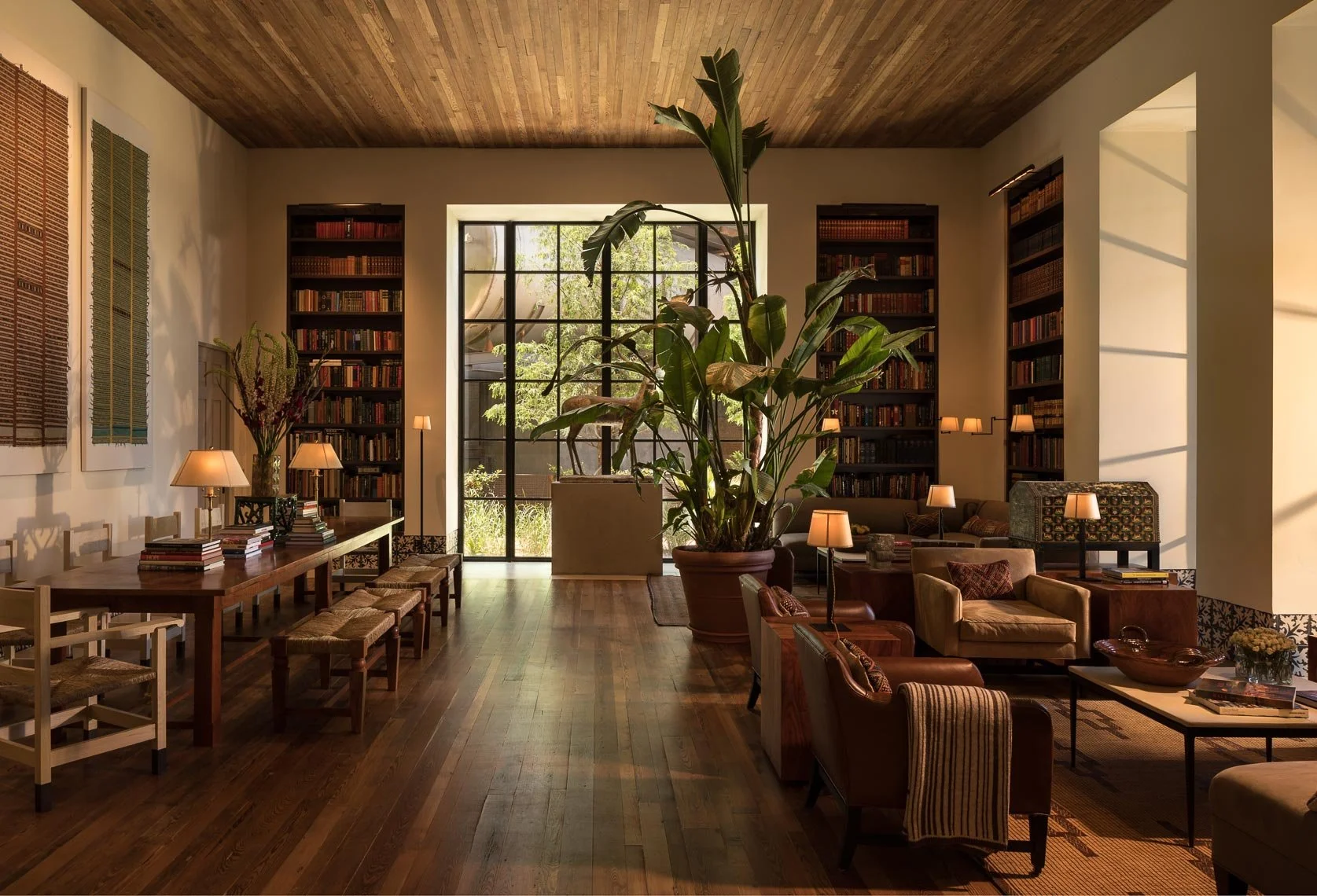 A cozy living room or library space with large windows, wooden floors, and wooden ceiling. The space features tall bookshelves filled with books, a long wooden table with chairs and books, a large potted plant, chairs and sofas with small lamps, and 