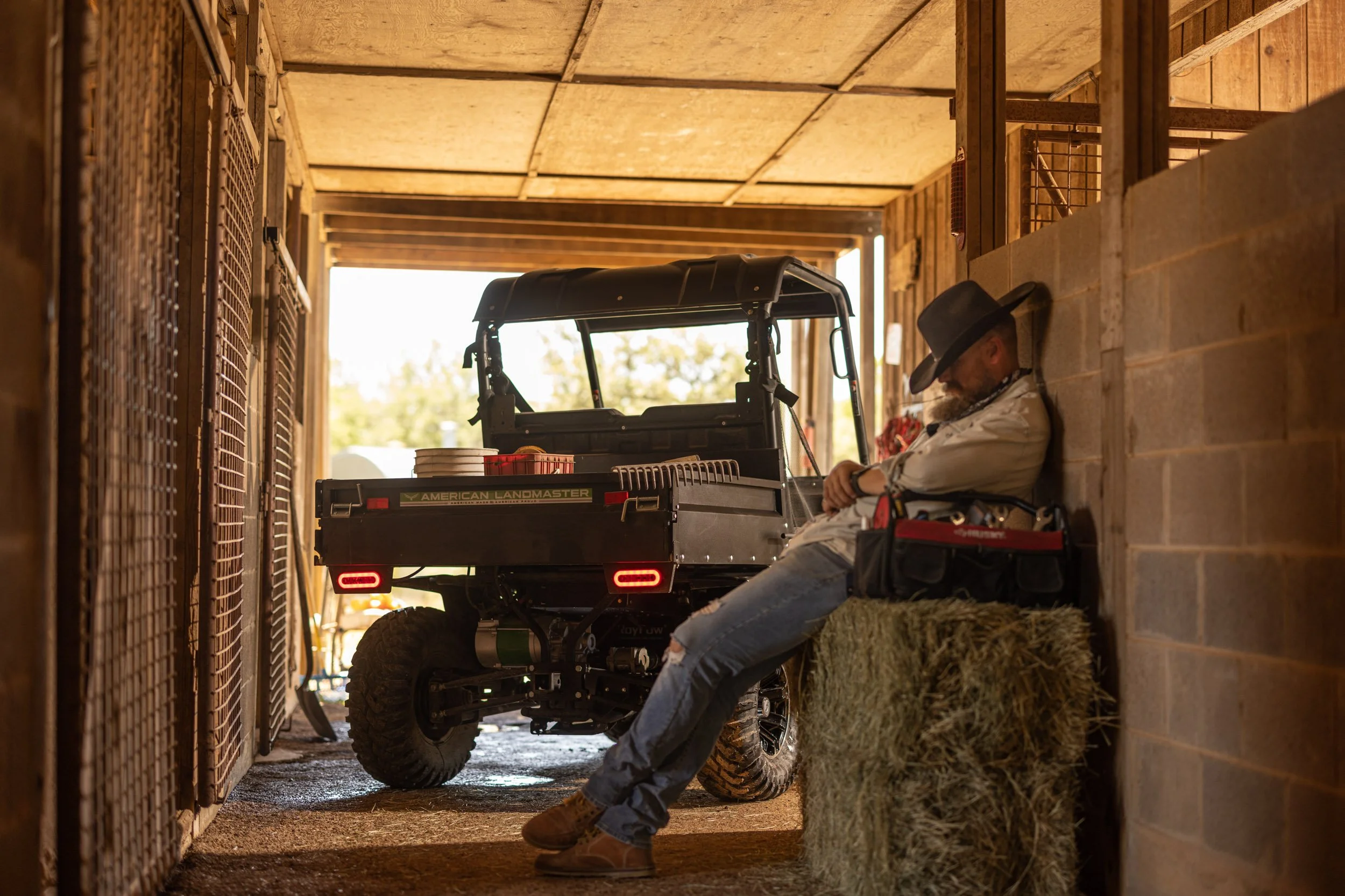 A man wearing a black hat and glasses, sitting with arms crossed, leaning against a hay bale in a wooden barn. Behind him a utility vehicle is parked near hay bales inside the barn.