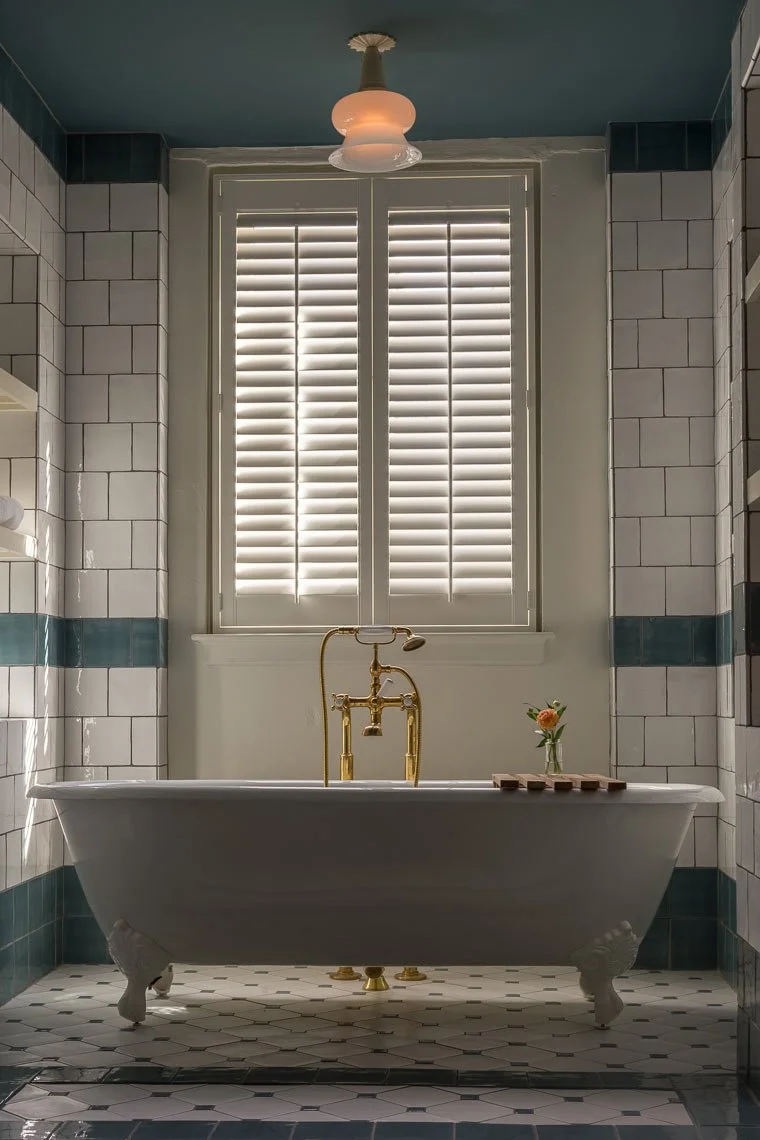 A vintage clawfoot bathtub with gold fixtures, a vase with a small flower, and a window with closed white shutters in a tiled bathroom.