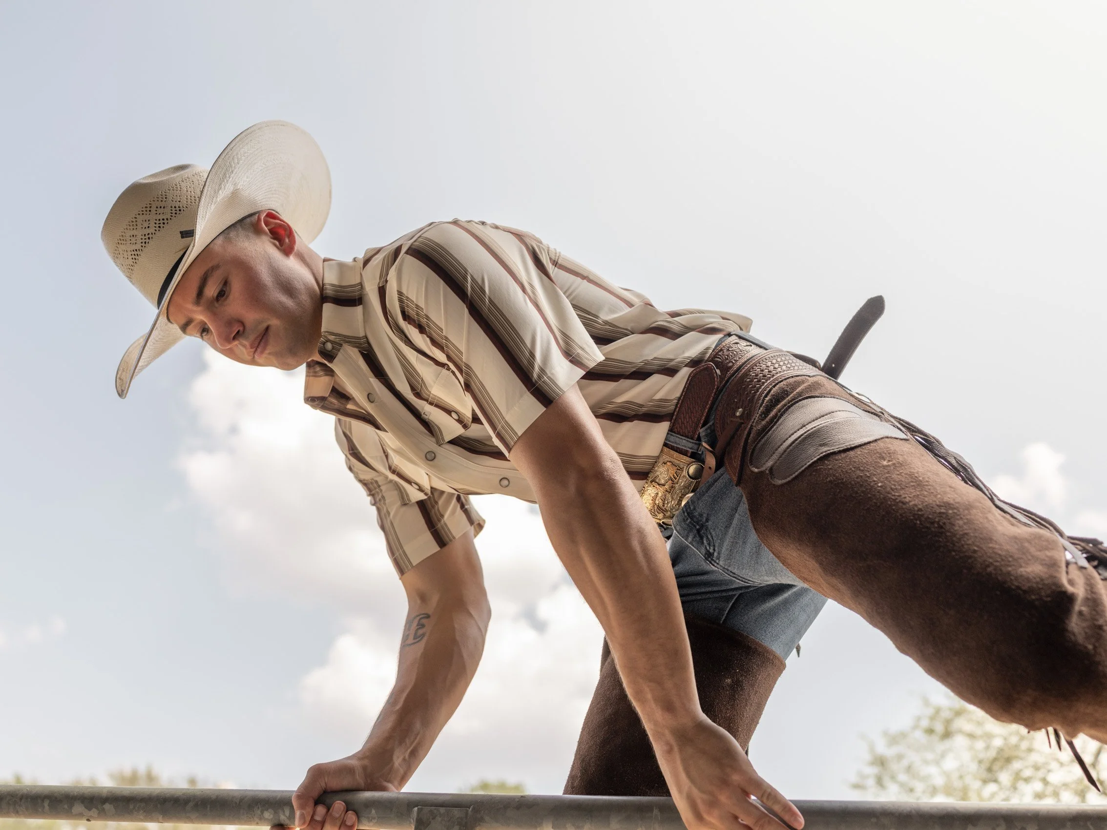 A young man wearing a cowboy hat, striped shirt, and shorts balancing on a metal railing outdoors during daylight, with a slightly cloudy sky in the background.