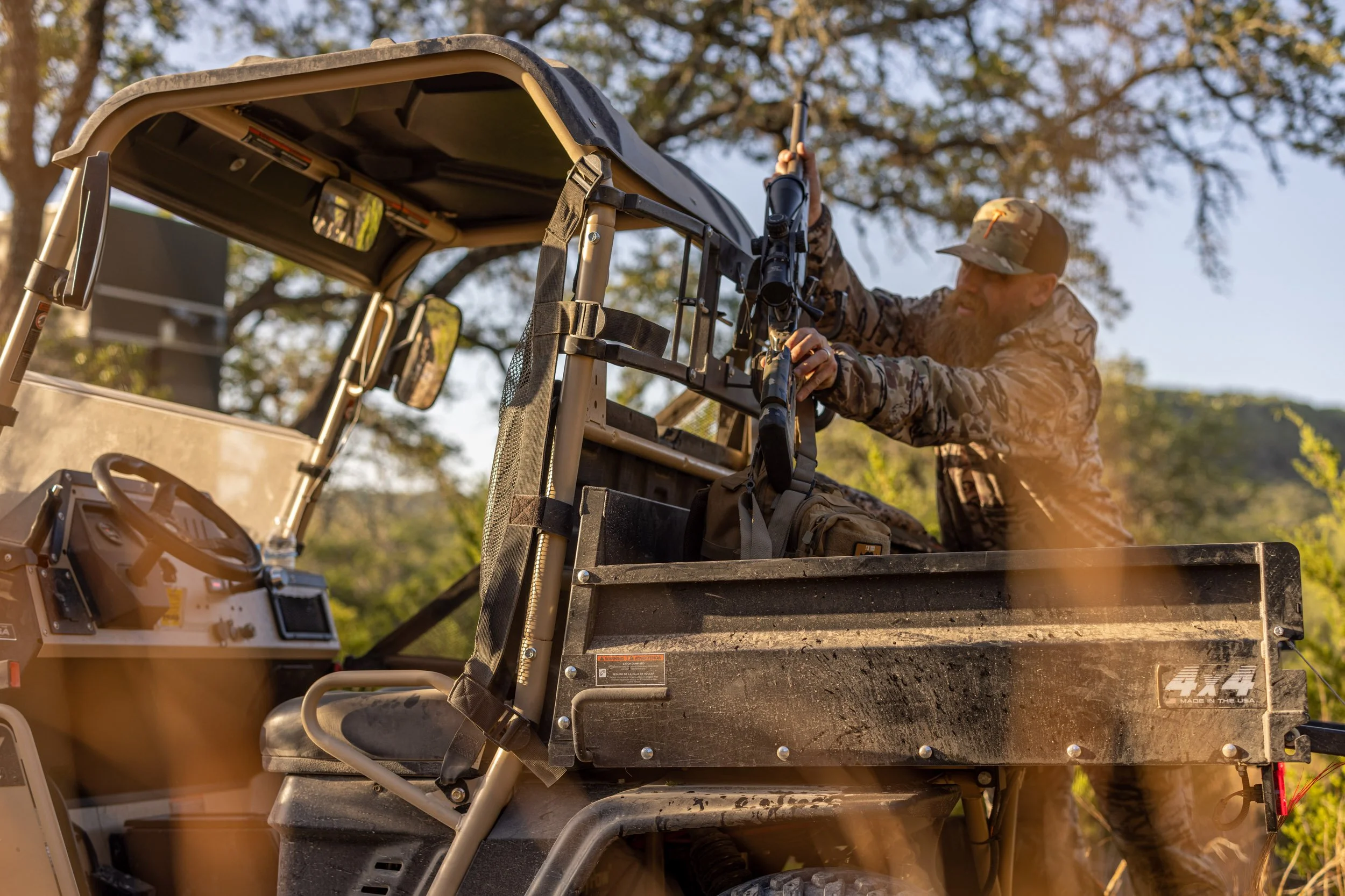 A man in camouflage clothing and cap aiming a scoped rifle from a hunting cart with a roof, on a sunny day with trees in the background.