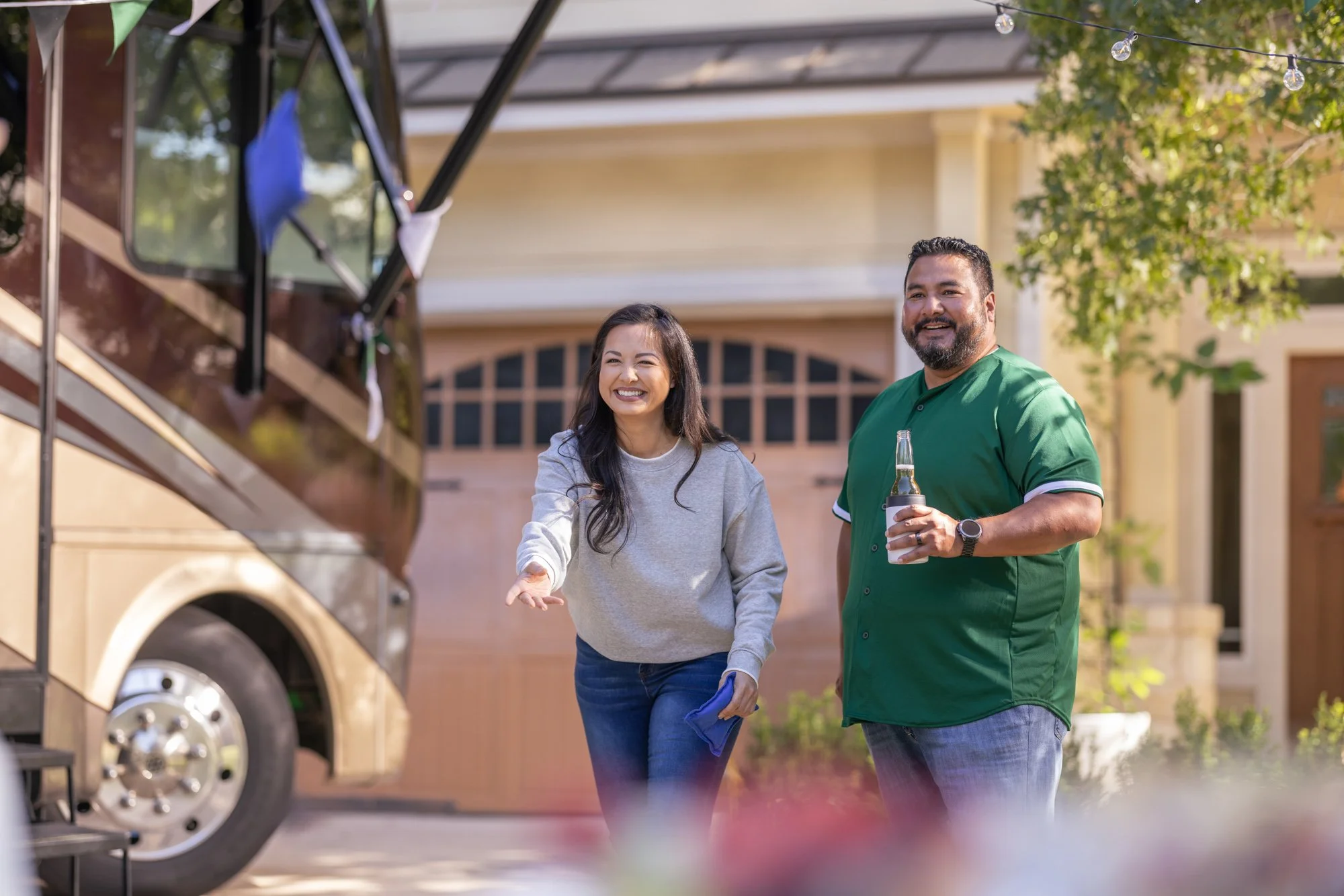 A woman and a man smiling and shaking hands outdoors near a food truck. The woman is wearing a grey sweatshirt and blue jeans, while the man is wearing a green shirt and jeans, holding a drink. The background includes a tree and a house.