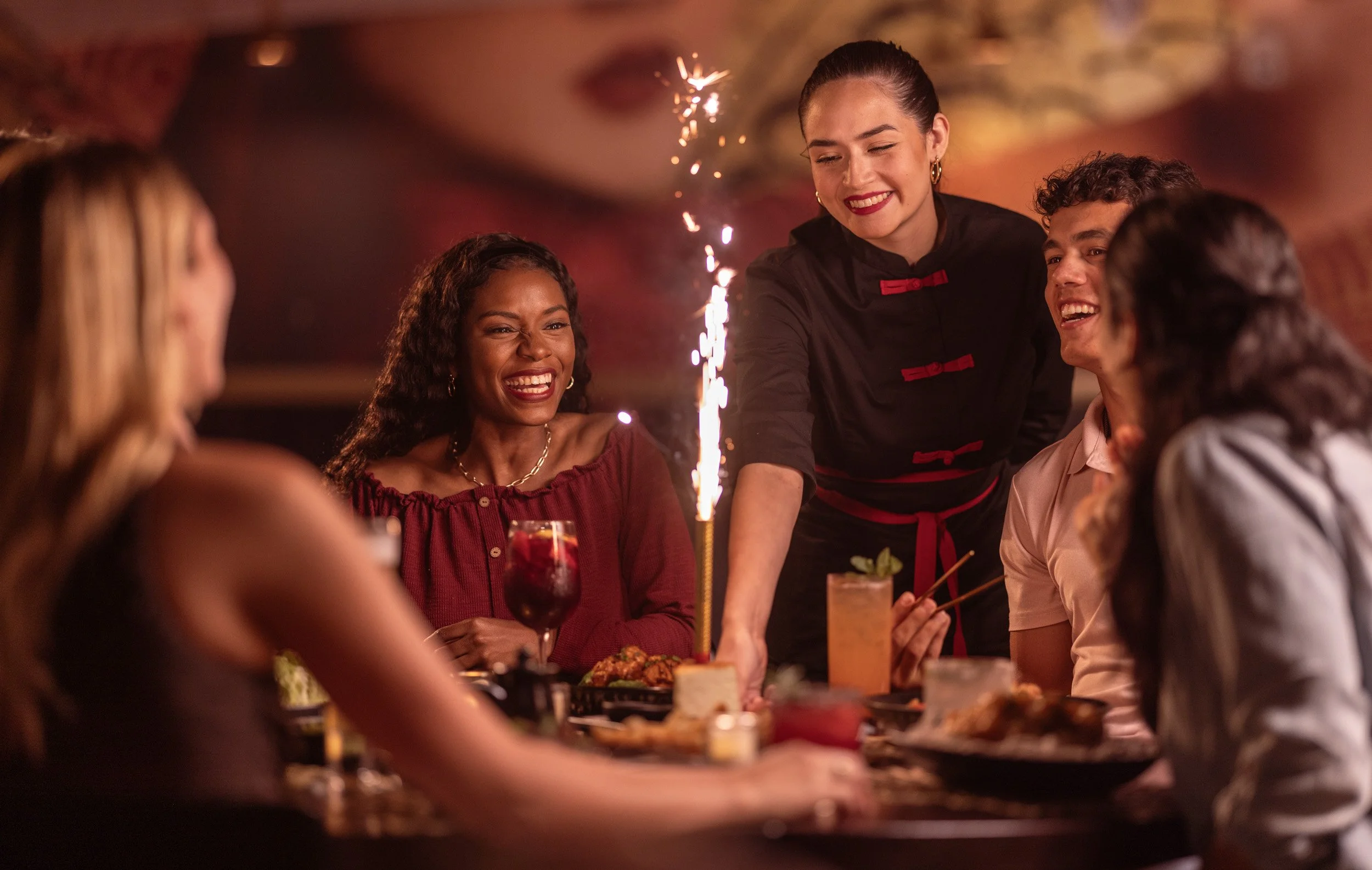 A group of diverse friends celebrating at a dinner table with a birthday cake that has a sparkler, with one person serving drinks, in a warm, lively atmosphere.