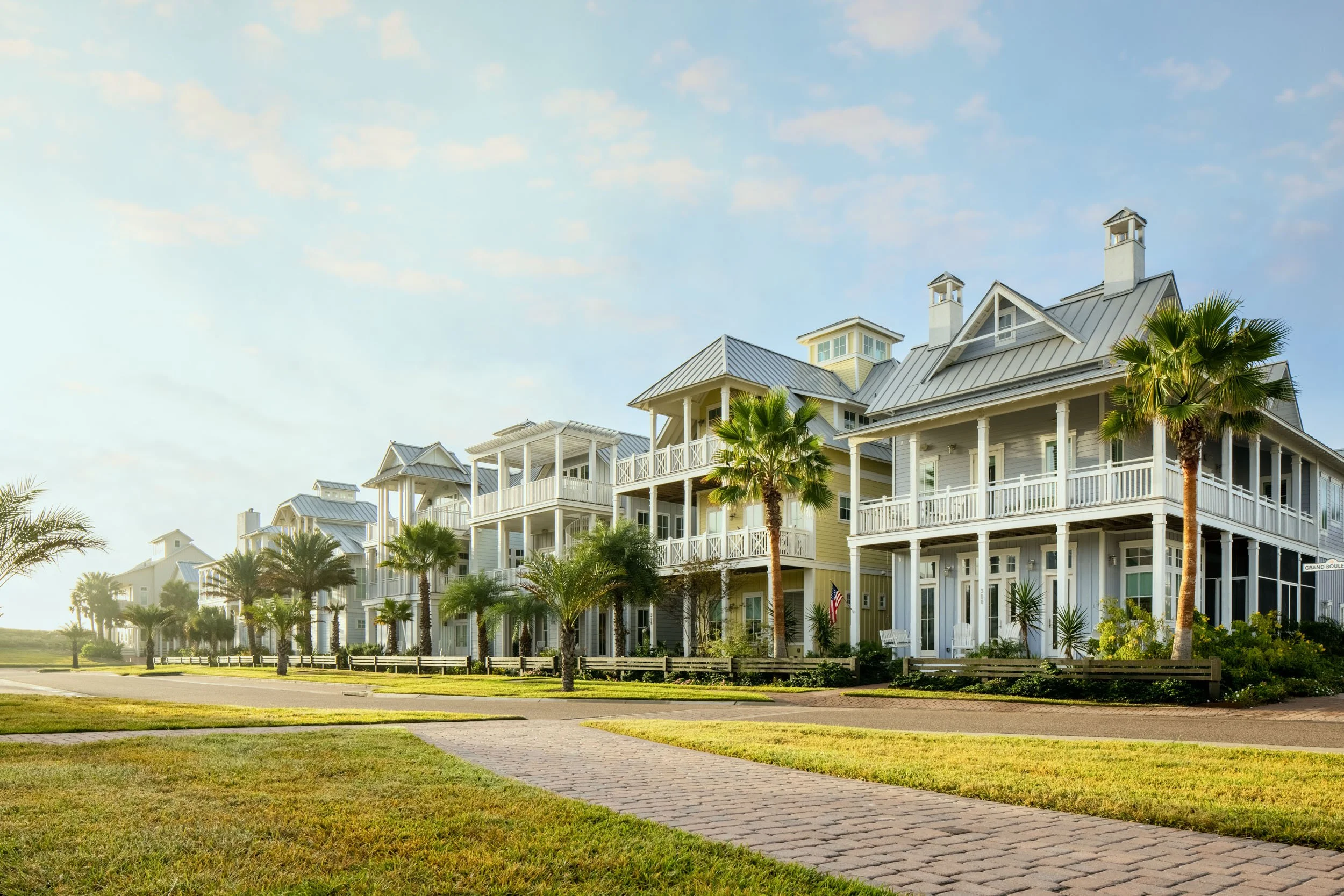 A row of large, white, colonial-style houses with wraparound porches, surrounded by palm trees, under a partly cloudy sky.