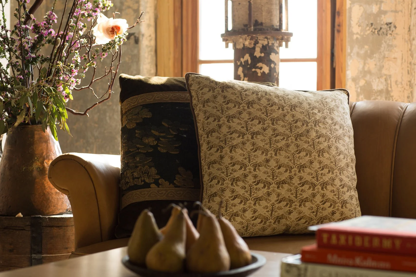 Close-up of a cozy living room scene featuring decorative pillows with animal and nature patterns on a beige sofa, a large vase with flowering branches and pink flowers, and a stack of books on a wooden table in front of the sofa, with a rustic indus