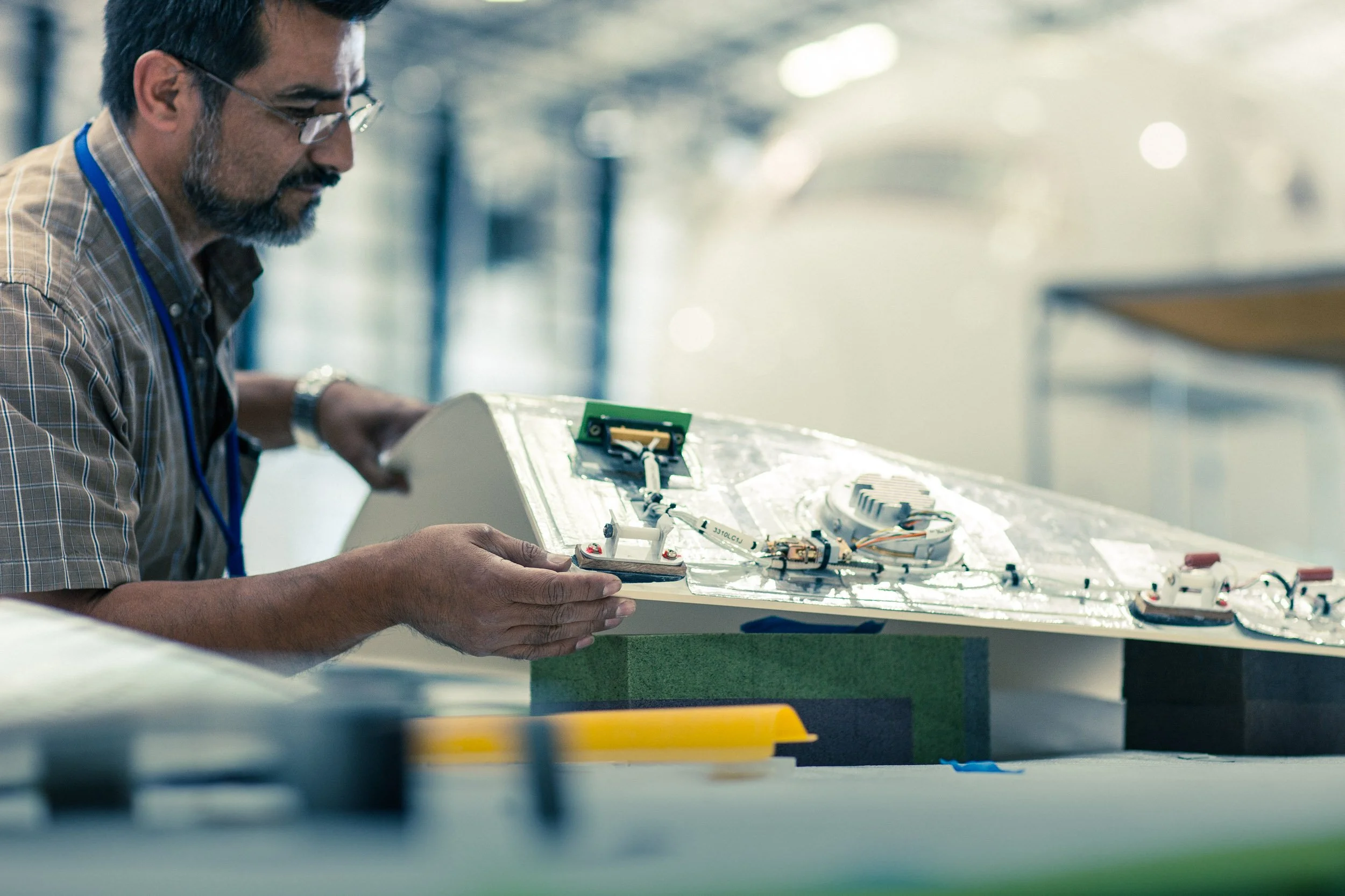 A man wearing glasses and a checkered shirt working on an electronic or mechanical project at a workbench in a well-lit workshop or laboratory.