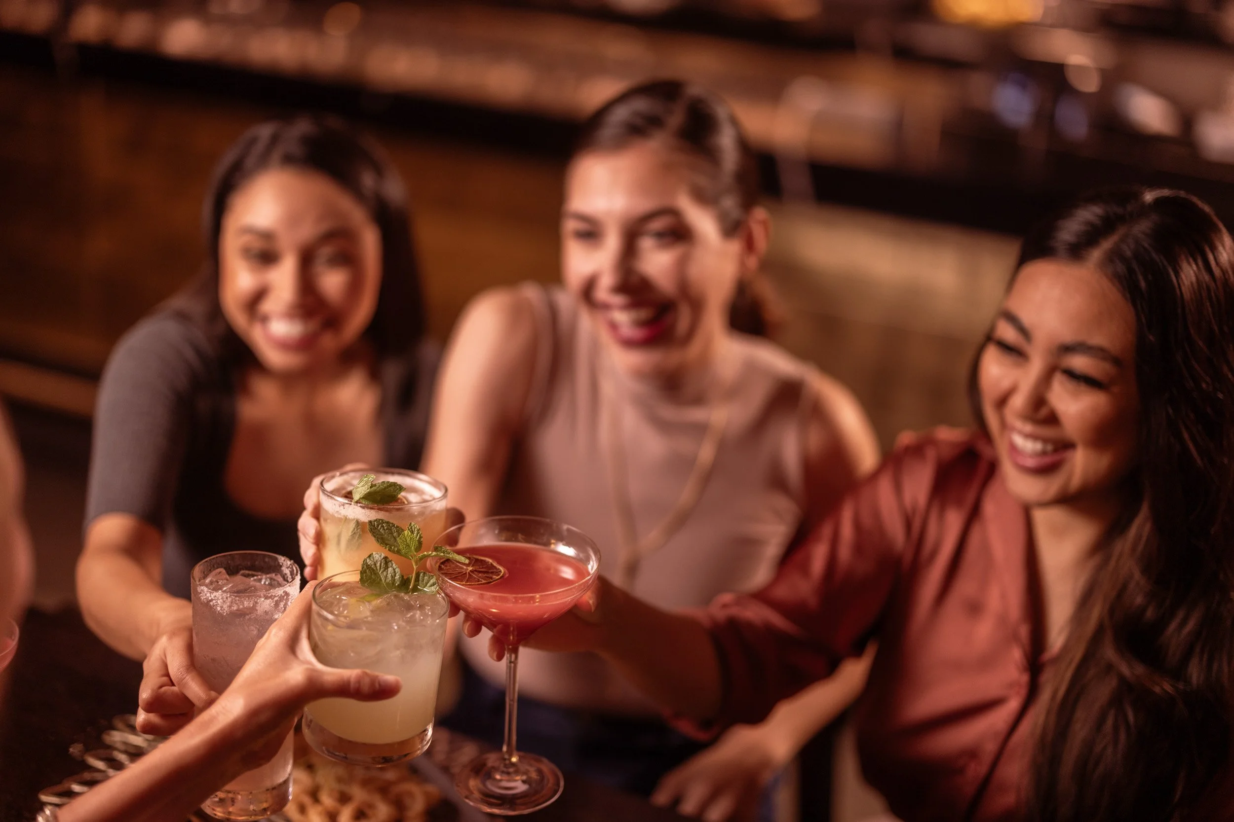 A group of friends at a bar raising their drinks in a toast, including a mojito, a margarita, and other cocktails, with three smiling women in the background.