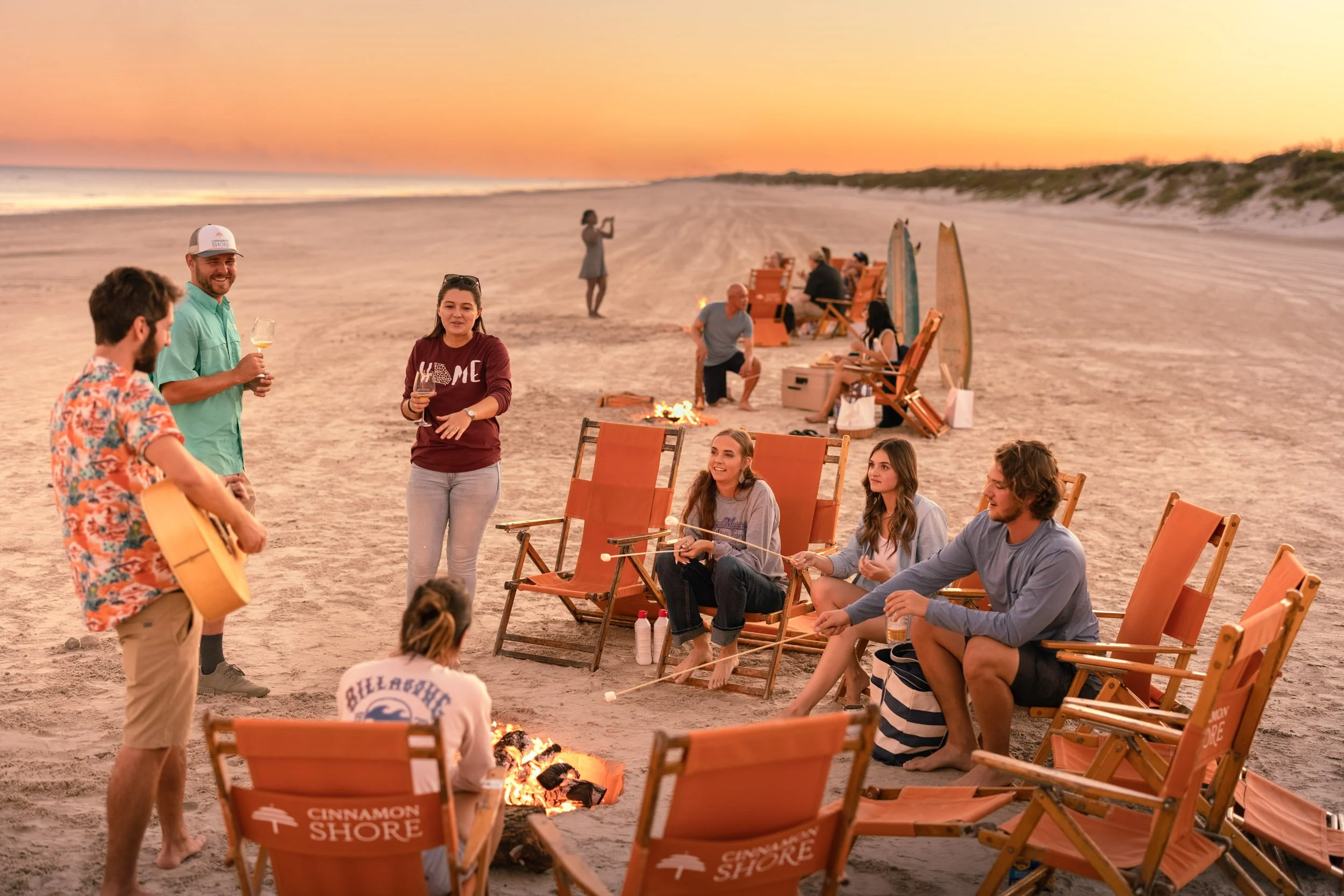 A group of people enjoying a beach bonfire sunset scene. Some are sitting on orange lounge chairs, others stand playing guitar and singing, with a fire pit in front. The background shows a sunset sky, ocean, and dunes.