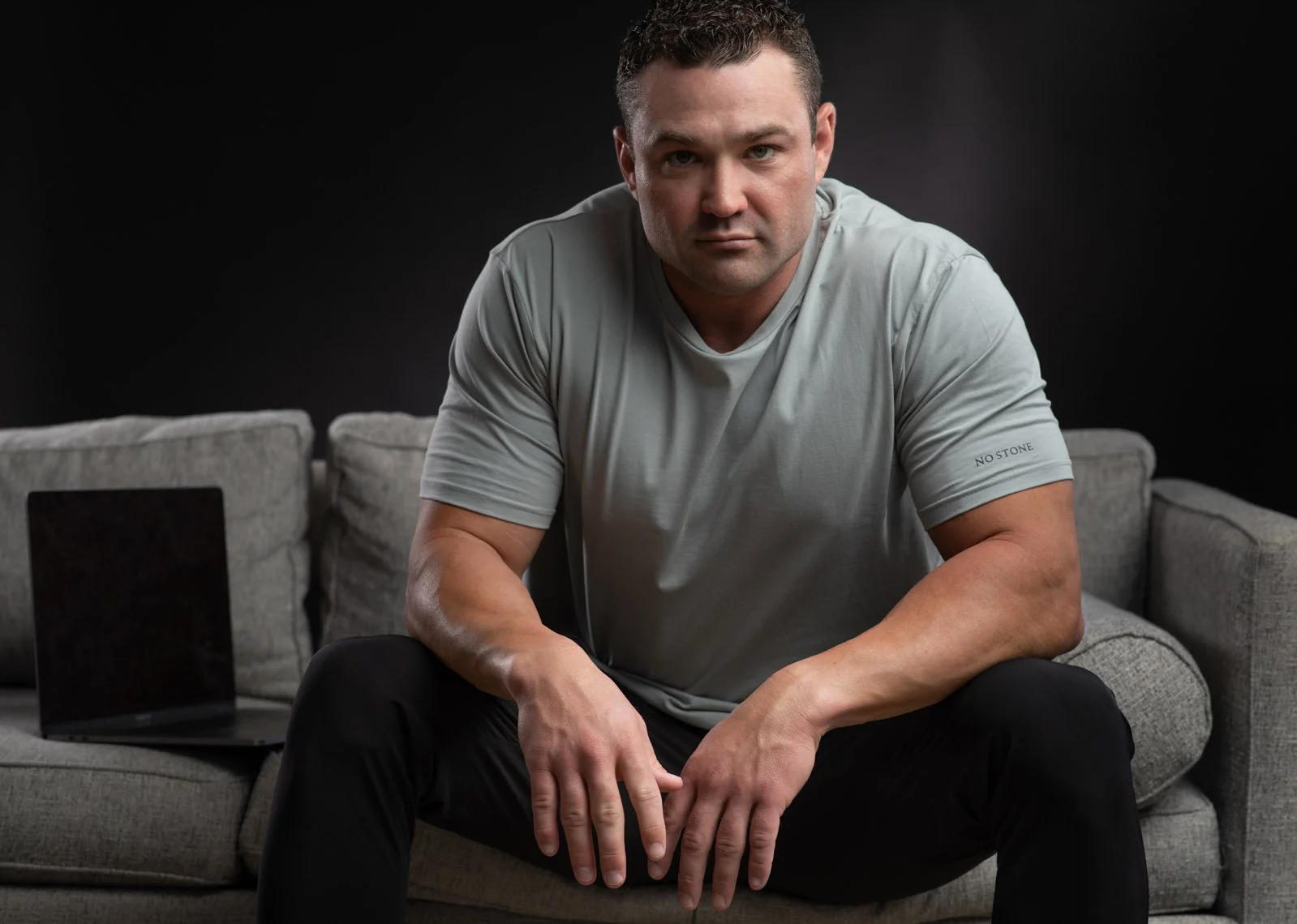 A man in a gray t-shirt sitting on a gray couch with a black background. A black laptop is open on the left side of the sofa.
