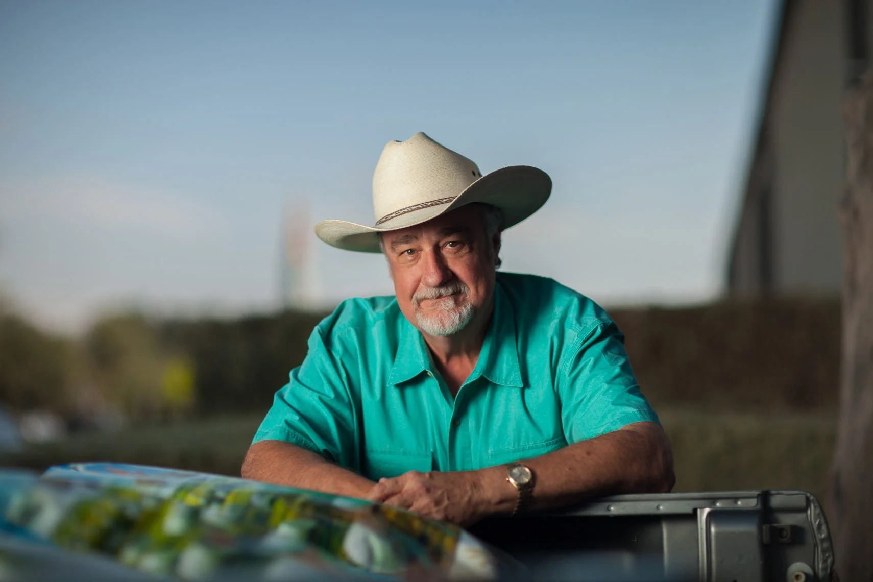 A man wearing a cowboy hat and a turquoise shirt sitting outdoors, looking at the camera with a serious expression, with a blue sky in the background.