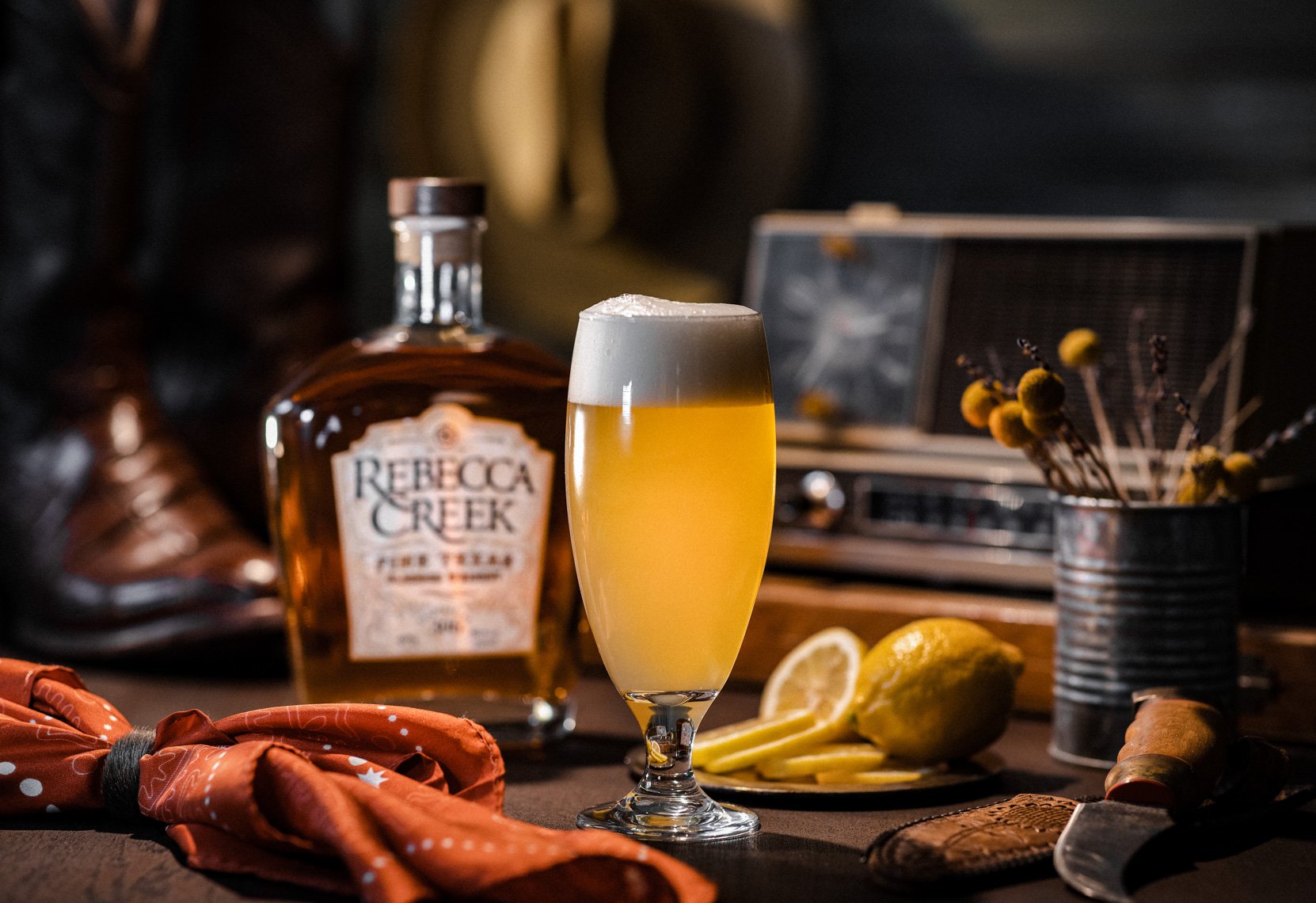 A glass of lemonade with lemon slices, a bottle of Rebecca Creek whiskey, a yellow lemon, a pair of sunglasses, a dried flower arrangement, and other objects on a dark wooden surface.