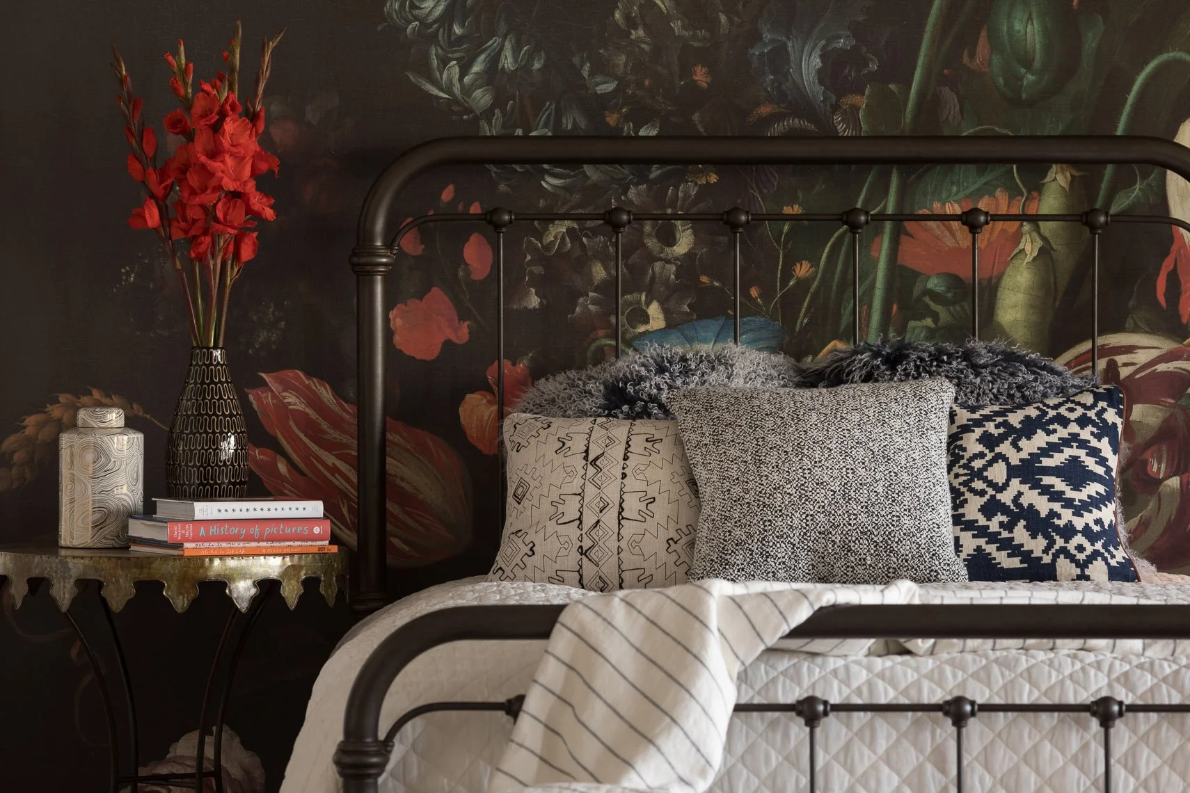 A bedroom with a black metal bed frame, decorated with various patterned pillows and a white quilted mattress. Behind the bed, there is a dark floral wallpaper with large red and green flowers. To the left of the bed, a gold side table holds a tall b