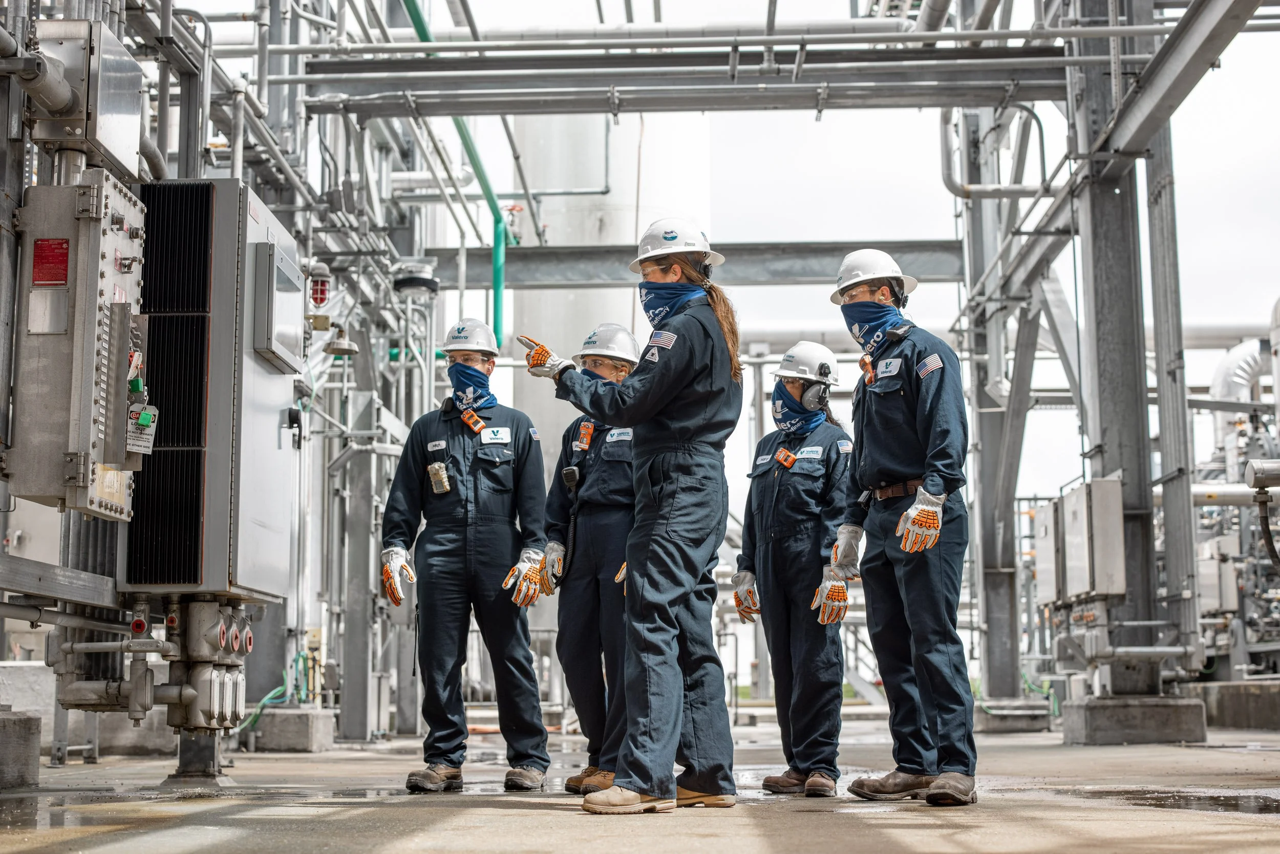 Five workers wearing navy uniforms, white helmets, and blue masks inspecting industrial equipment in an outdoor facility with metal piping and frameworks.
