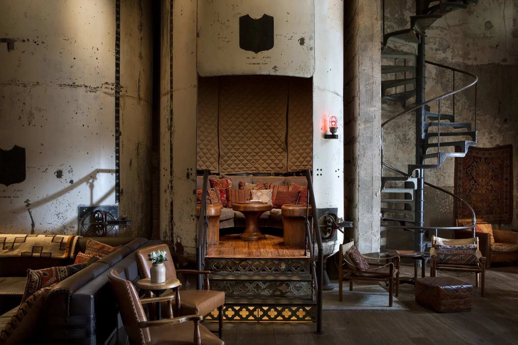 Interior of a vintage-style lounge with industrial elements: a cozy seating area with brown leather armchairs, patterned cushions, a wooden table, a spiral staircase, and exposed concrete walls with patches of paint.