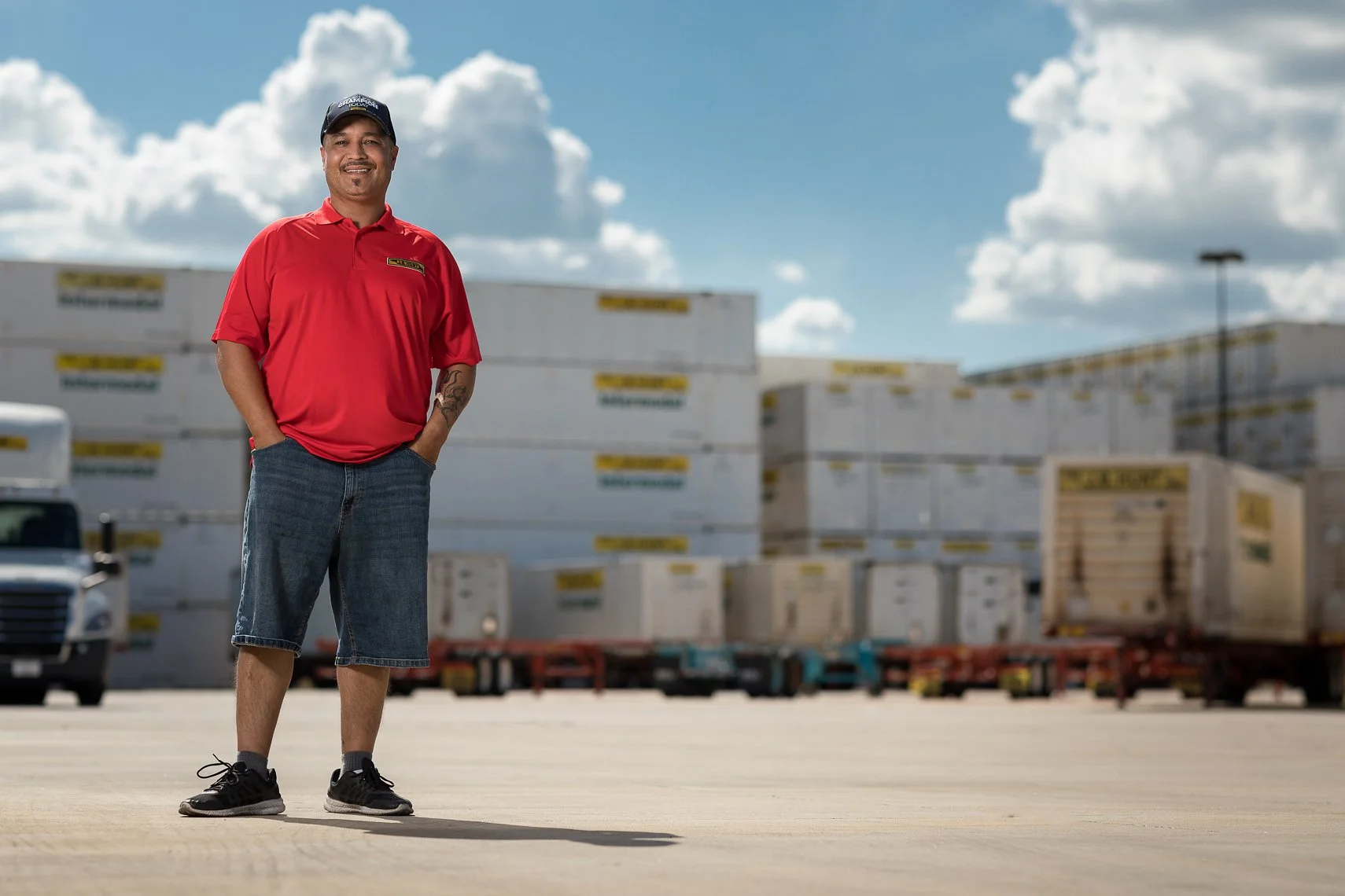 A man wearing a red polo shirt, denim shorts, and black sneakers standing in an outdoor industrial area with stacked shipping containers and a truck in the background.