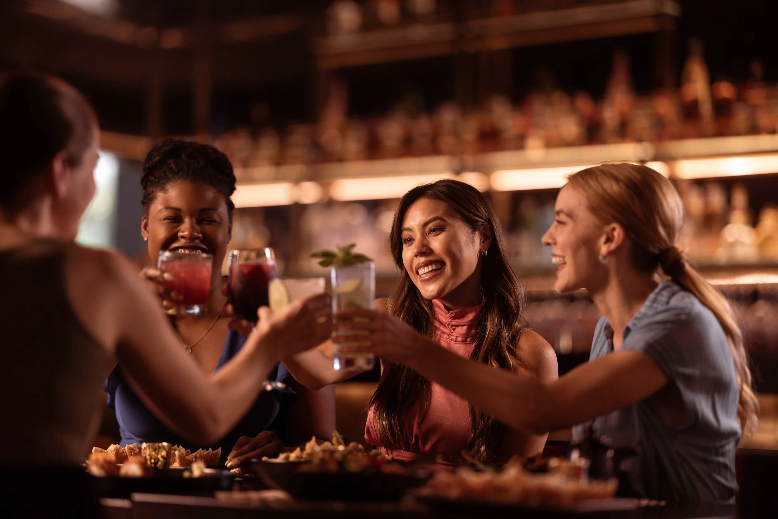 Group of friends at a bar raising glasses in a toast, smiling and enjoying drinks in a dimly lit setting.
