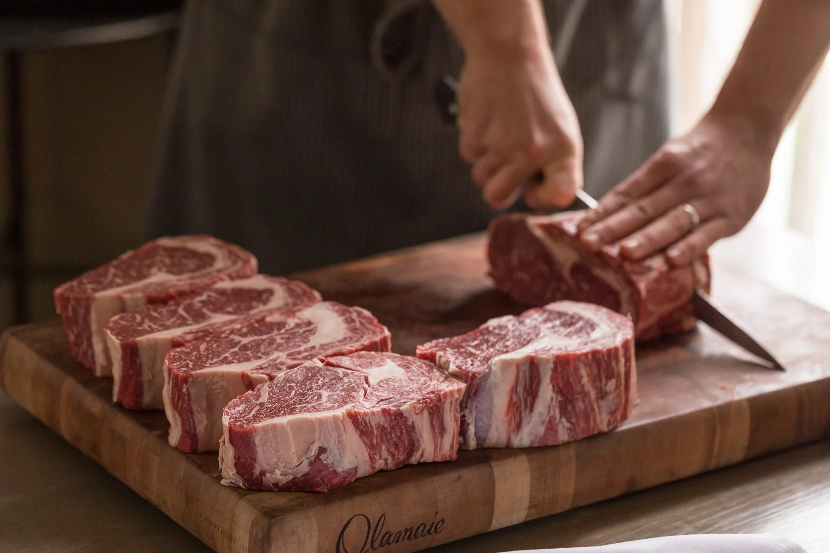 Person slicing raw beef steaks on a wooden cutting board with additional steaks laid out.
