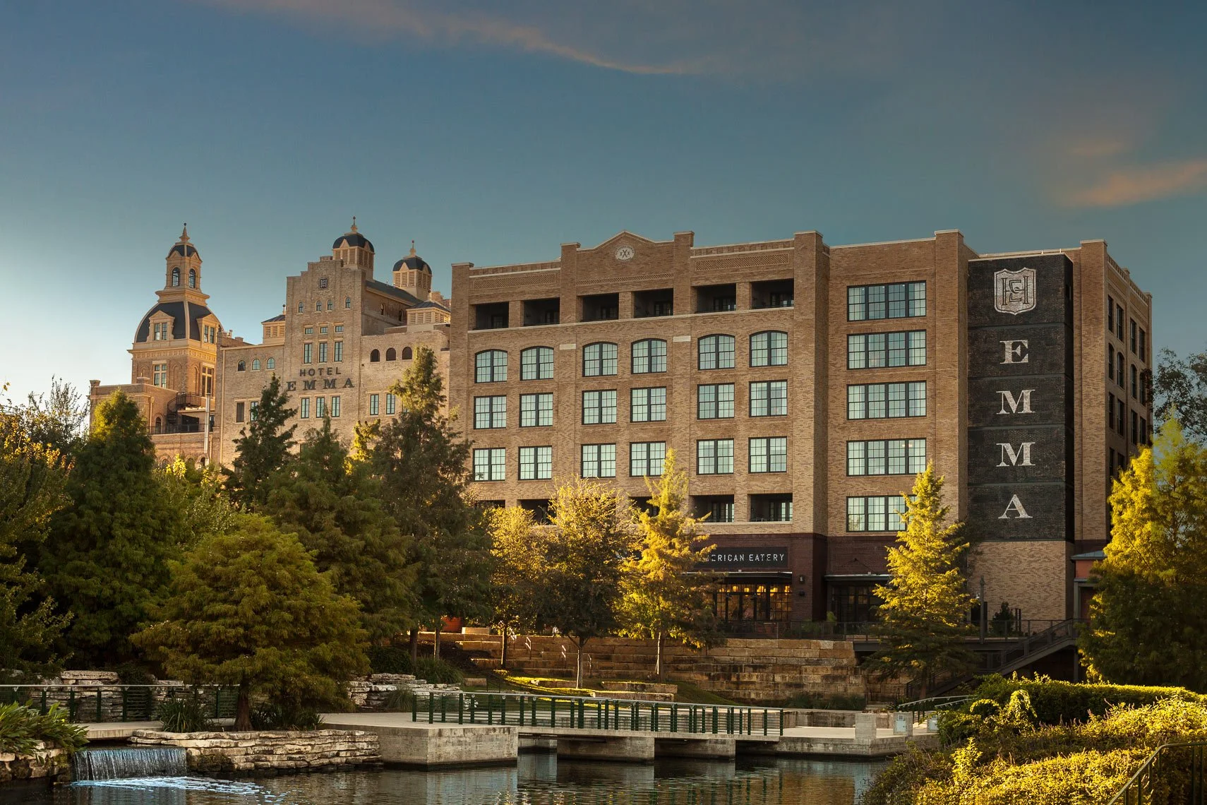 A multi-story brick hotel named Hotel Emma with towers and domes, surrounded by trees and a small water feature in the foreground, with a clear sky at sunset.