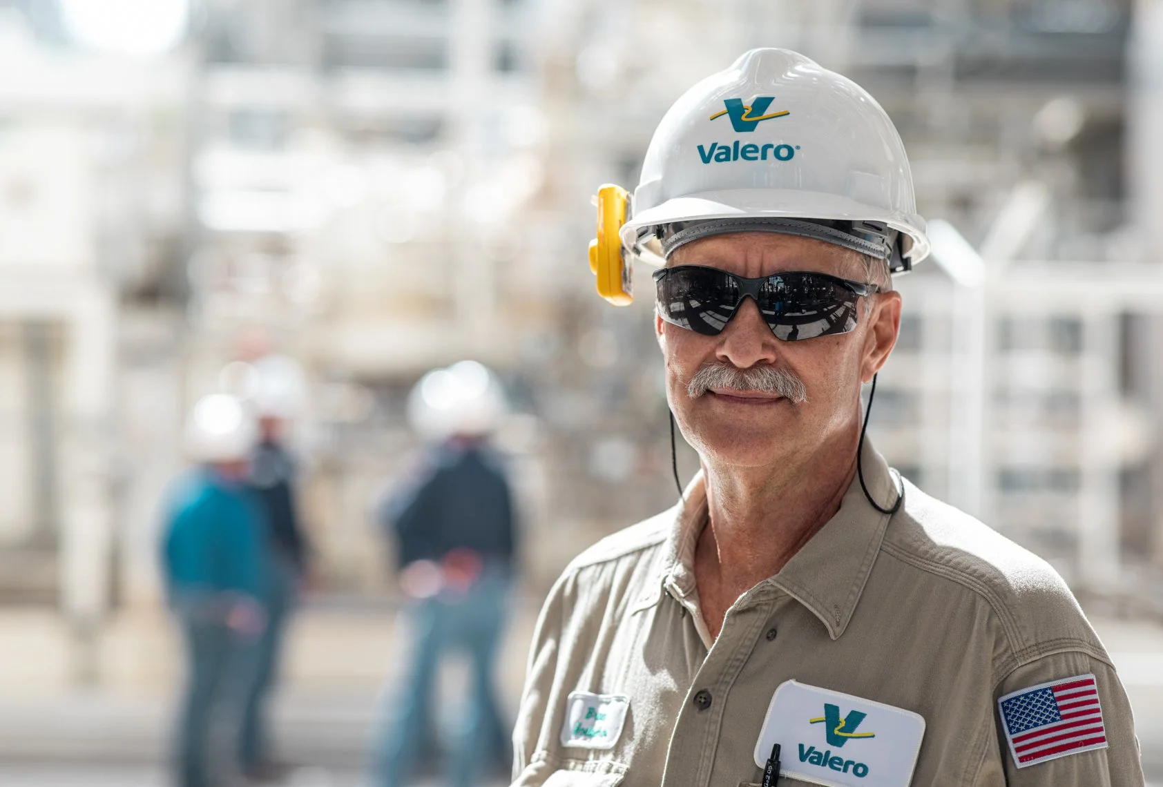 A man wearing sunglasses, a Valero hard hat, and a security badge in an industrial setting with blurred workers and equipment in the background.