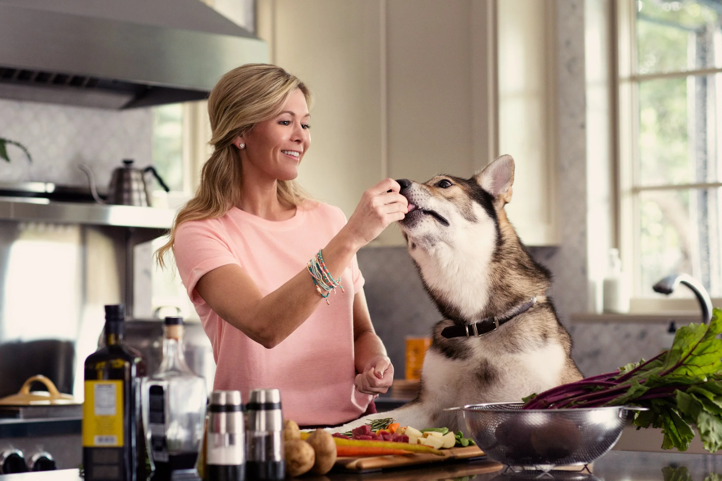 A woman in a pink shirt feeding a large husky dog in a kitchen with large windows.
