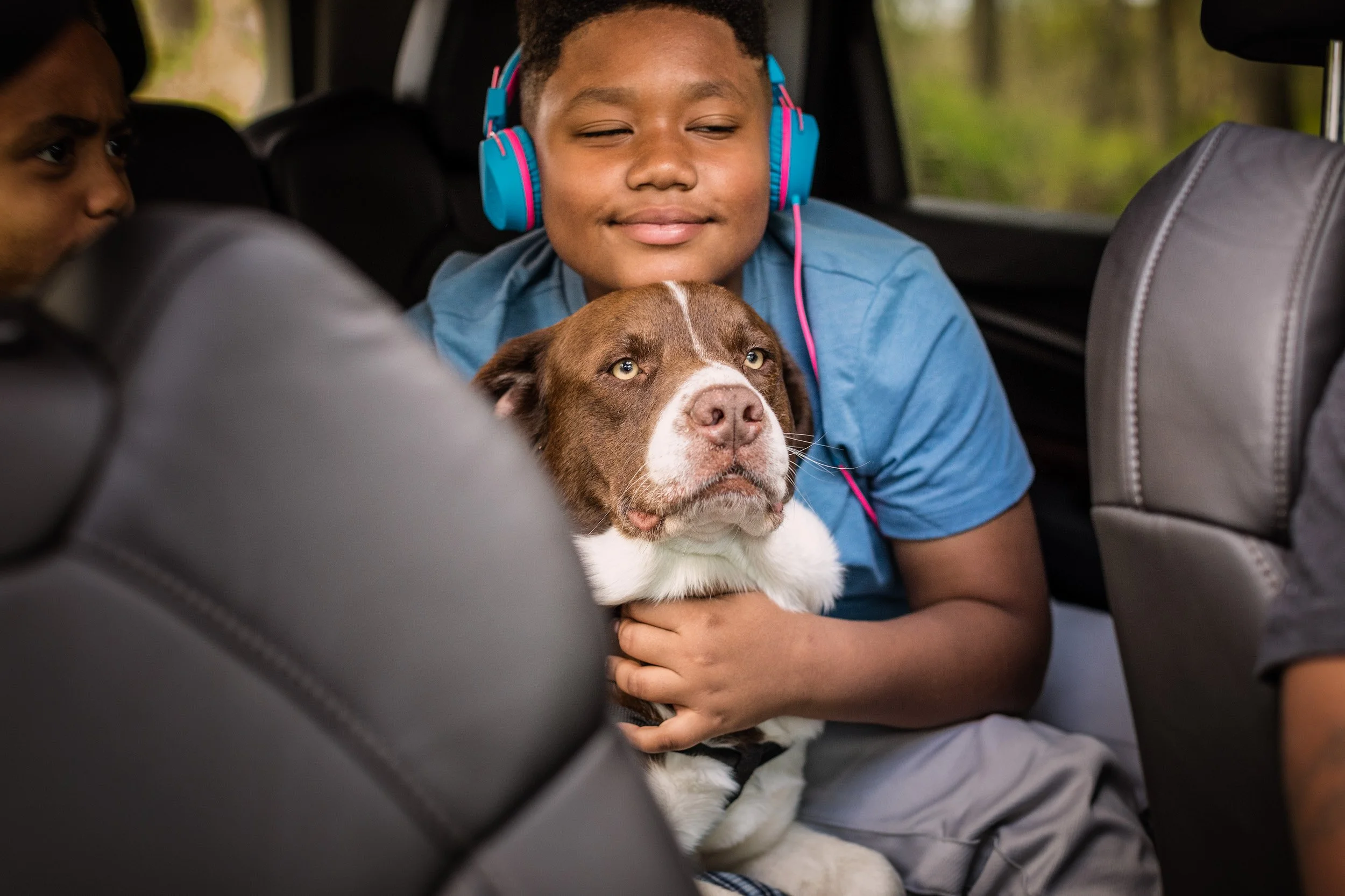 A boy with blue headphones holding a brown and white dog in the back seat of a vehicle.