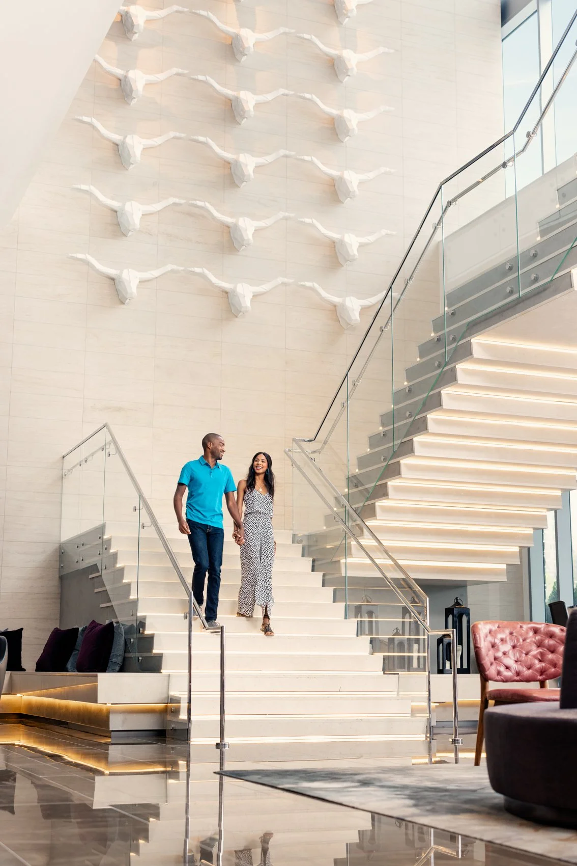 A man and woman walking down a staircase inside a modern building, holding hands and smiling. The staircase has glass railings and a decorative wall sculpture of several white abstract figures on the beige wall behind them.