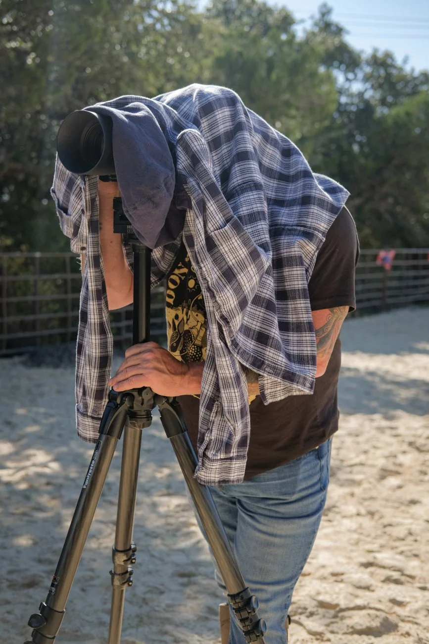 Person in black T-shirt and blue jeans looking through a telescope on a sandy ground outdoors.