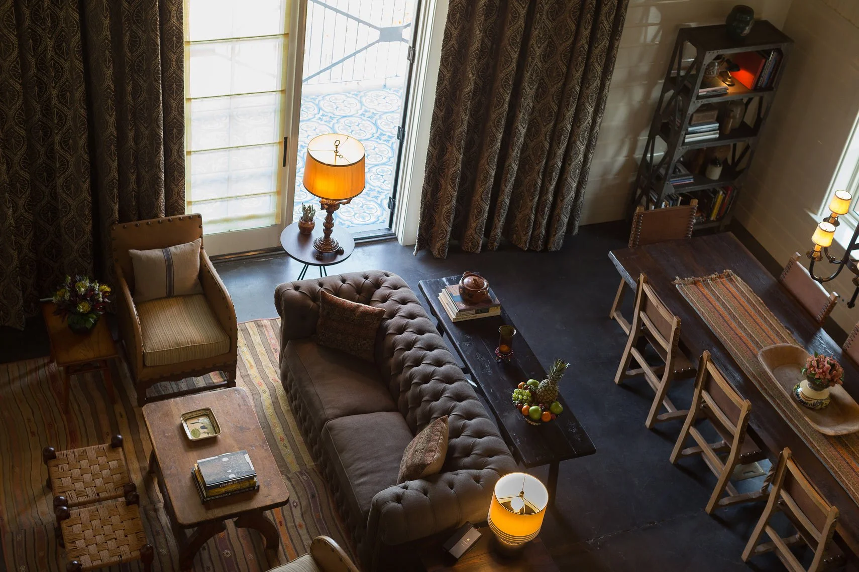 A cozy living room with a sliding glass door leading to an outdoor area with patterned tiles. The room features a beige armchair with a pillow, a brown tufted sofa with a throw pillow, a wooden coffee table with books, a side table with a lamp, a bla