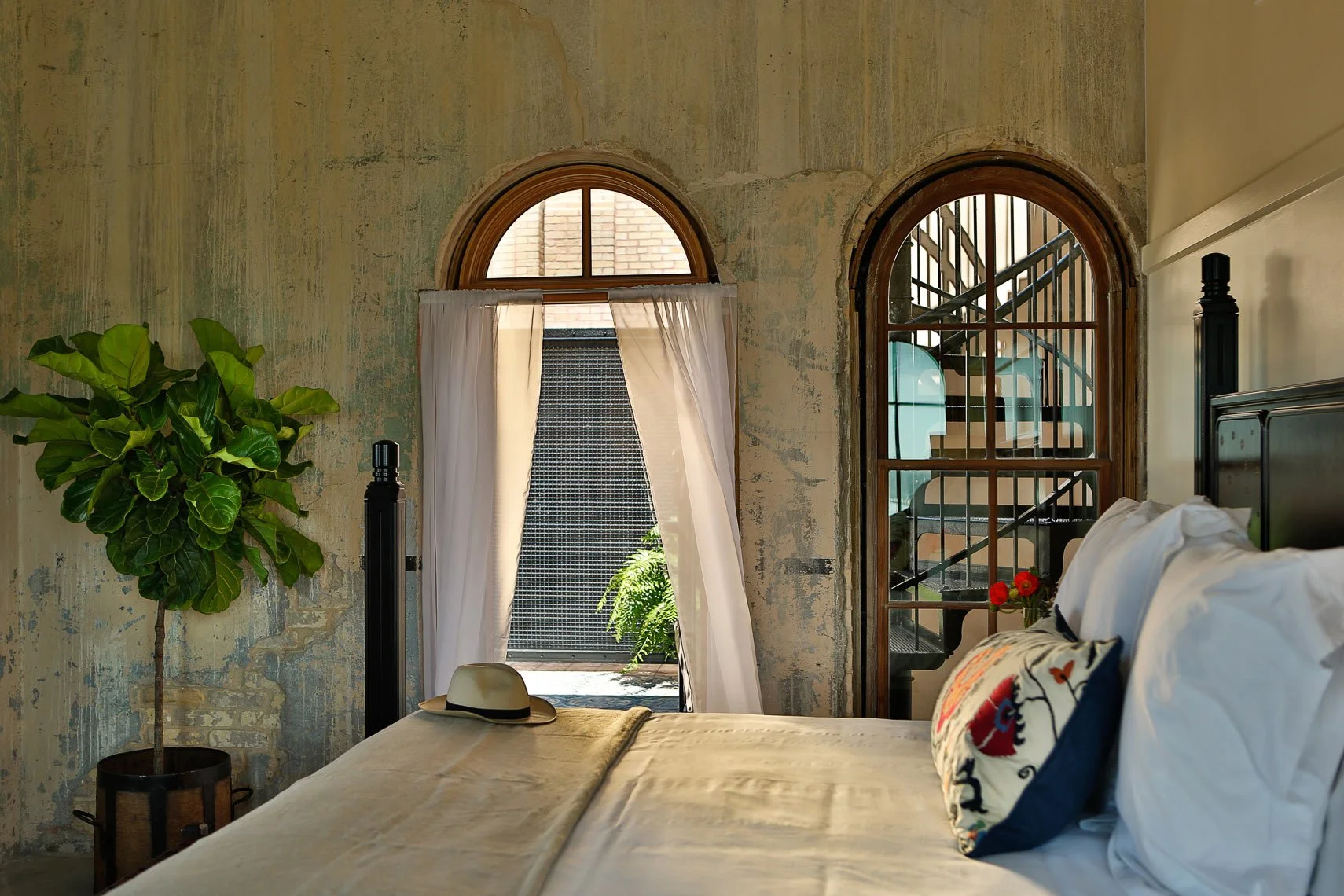 A bedroom with a bed, pillows, a hat on the bed, a large green potted plant, and two arched windows with curtains. There is a staircase visible through the right window, suggesting an industrial or loft-style space.