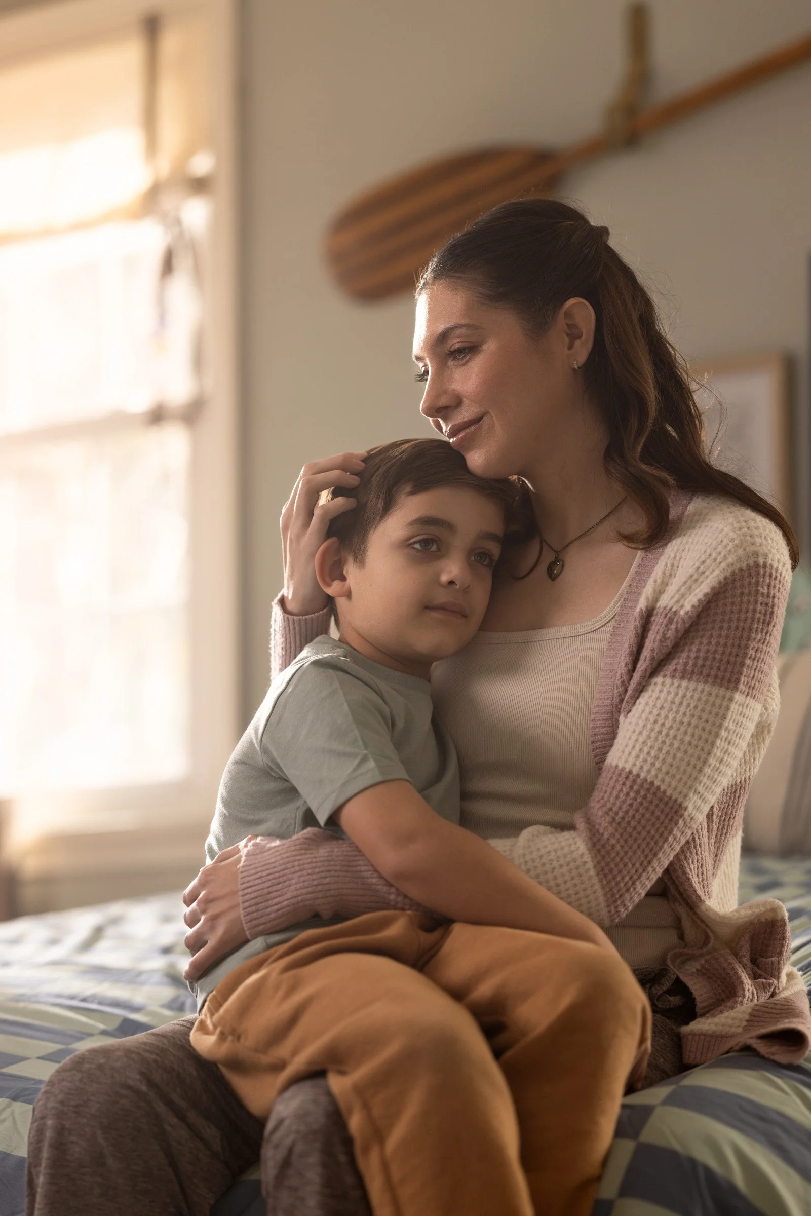 A woman with long, brown hair and a boy with short dark hair are sitting on a bed near a window, embracing, with the woman gently touching the boy's head and smiling softly.