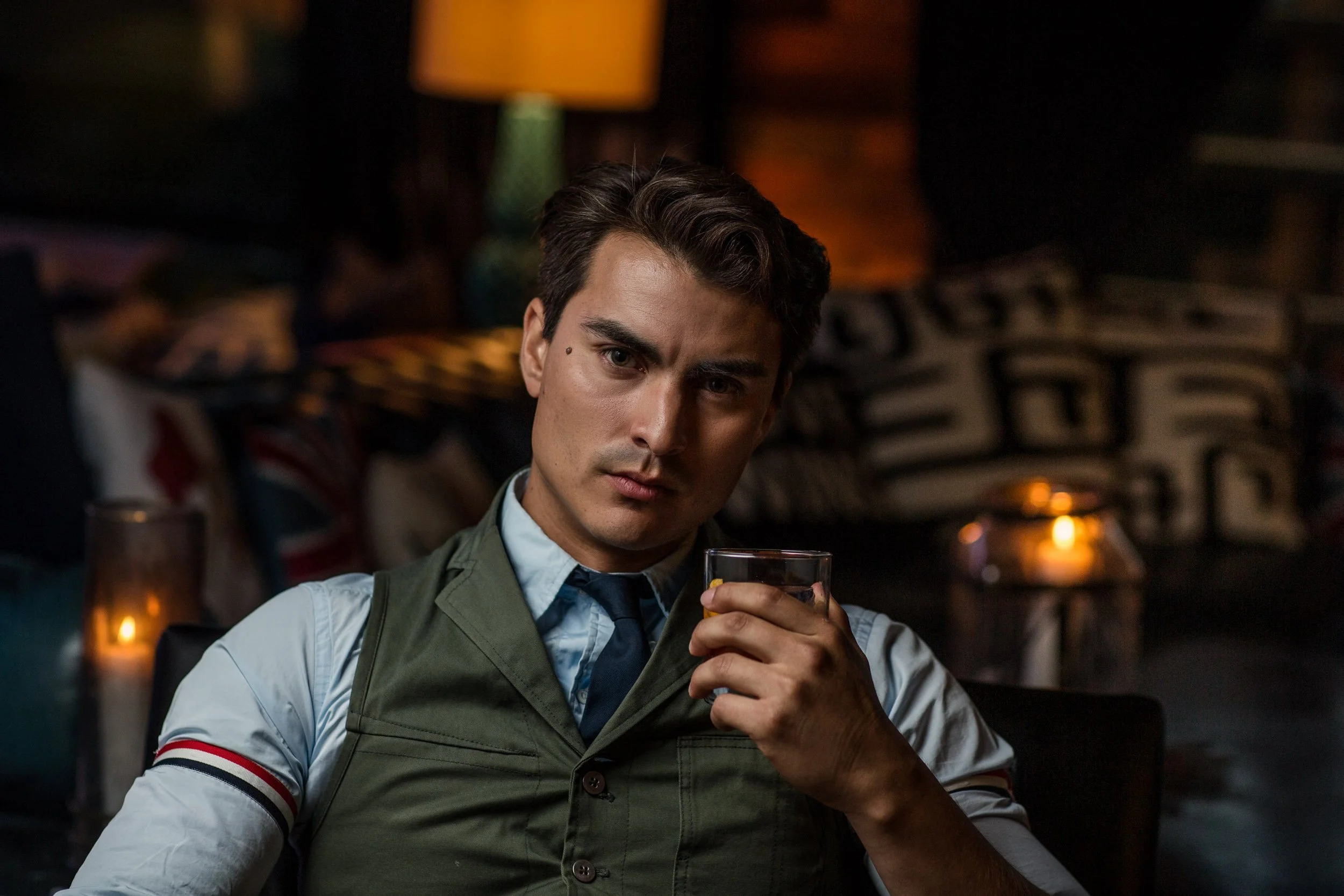 A young man with dark hair wearing a gray vest over a white shirt, holding a glass of drink, sitting in a dimly lit room with warm lighting and decorative cushions in the background.