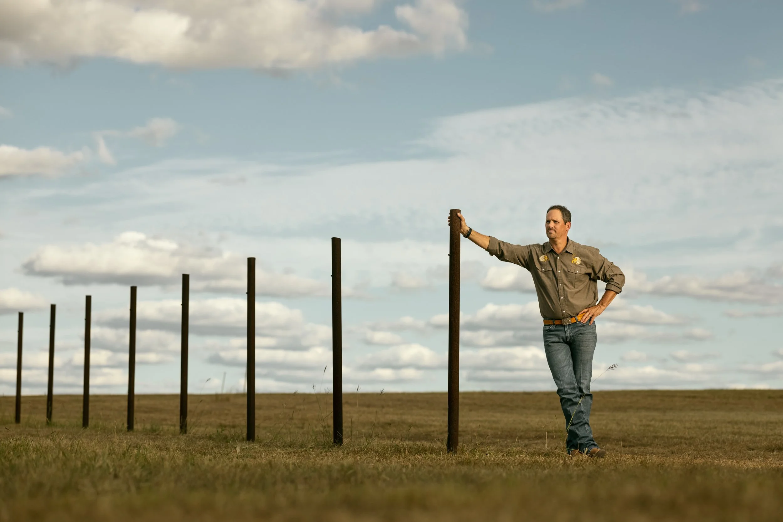 A man stands outdoors next to a row of vertical metal posts, with his right arm resting on one of them. He is wearing a tan shirt with patches and dark jeans, looking to his left with a serious expression. The sky is partly cloudy with blue and white