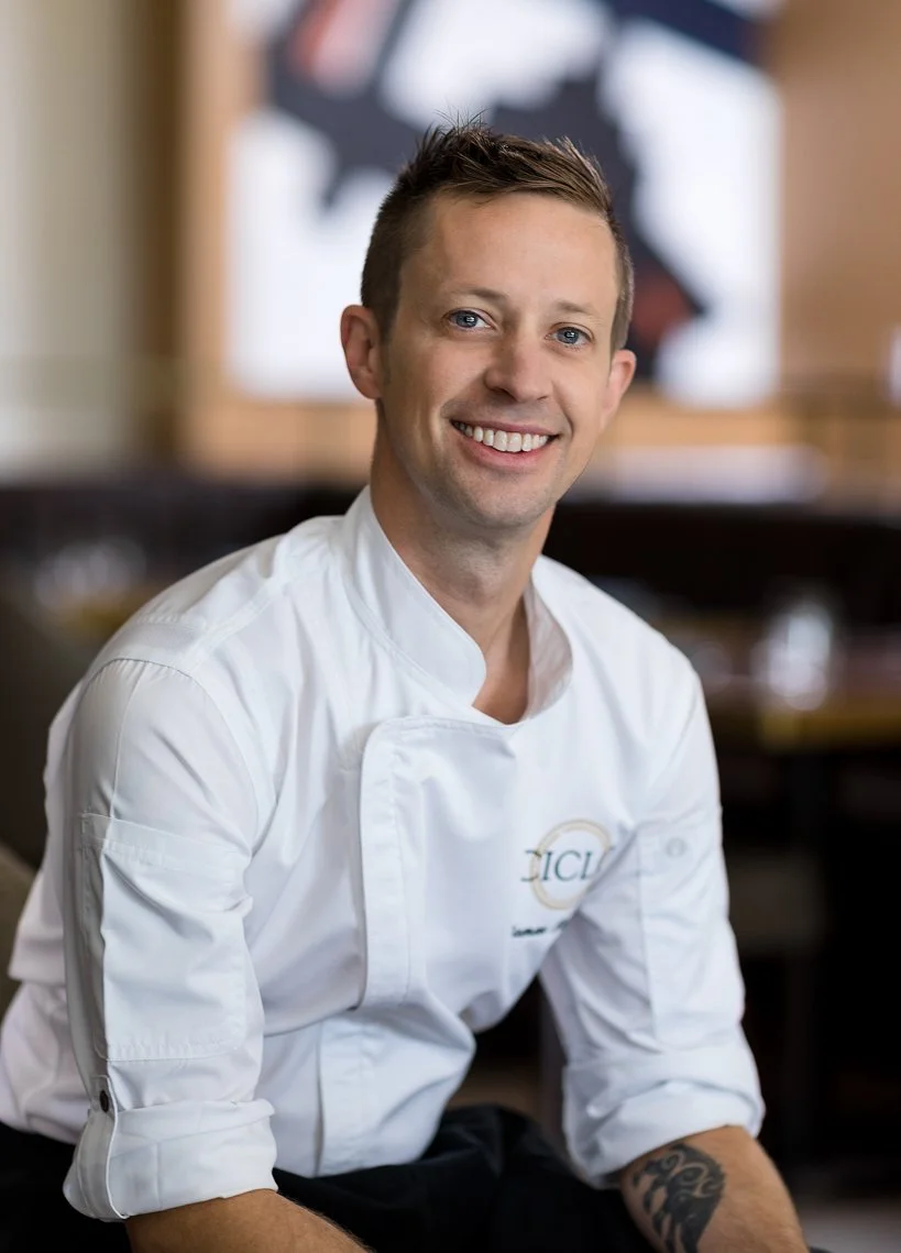 A young man with short brown hair and blue eyes is smiling. He is wearing a white chef's jacket with a logo, and has a tattoo on his left forearm. The background is blurred but appears to be a culinary or restaurant setting.