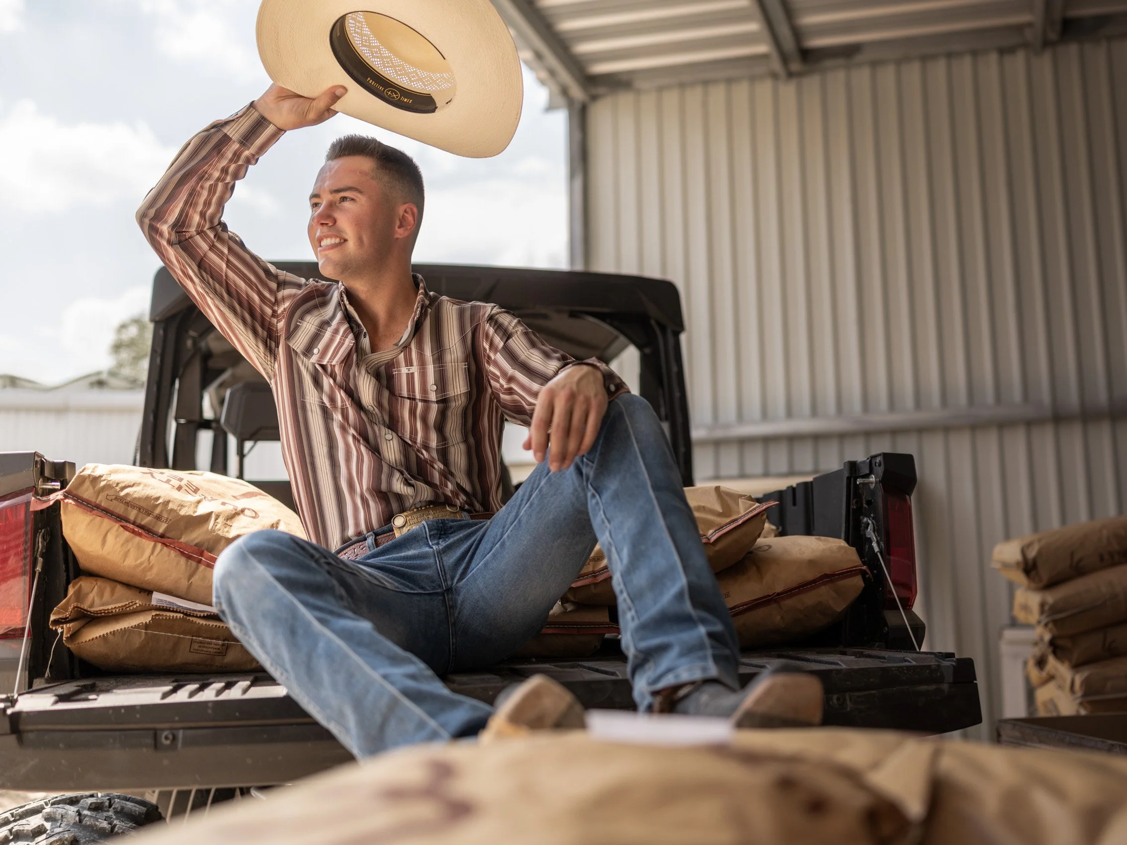 A young man with short hair, wearing a plaid shirt and jeans, sitting on the back of a pickup truck inside a barn, holding a straw cowboy hat above his head and smiling.