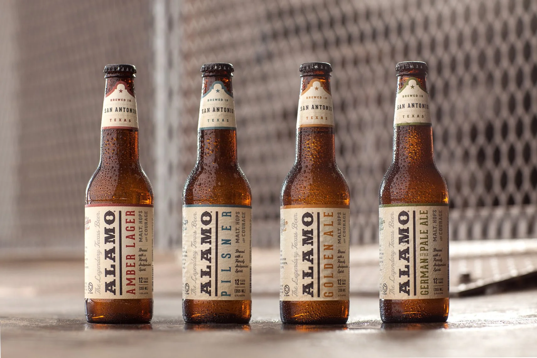 Four bottles of beer with condensation, labeled from left to right as Amber Lager, Paslne, Golden Ale, and German Pale Ale, set on a wooden surface with a blurred metallic background.