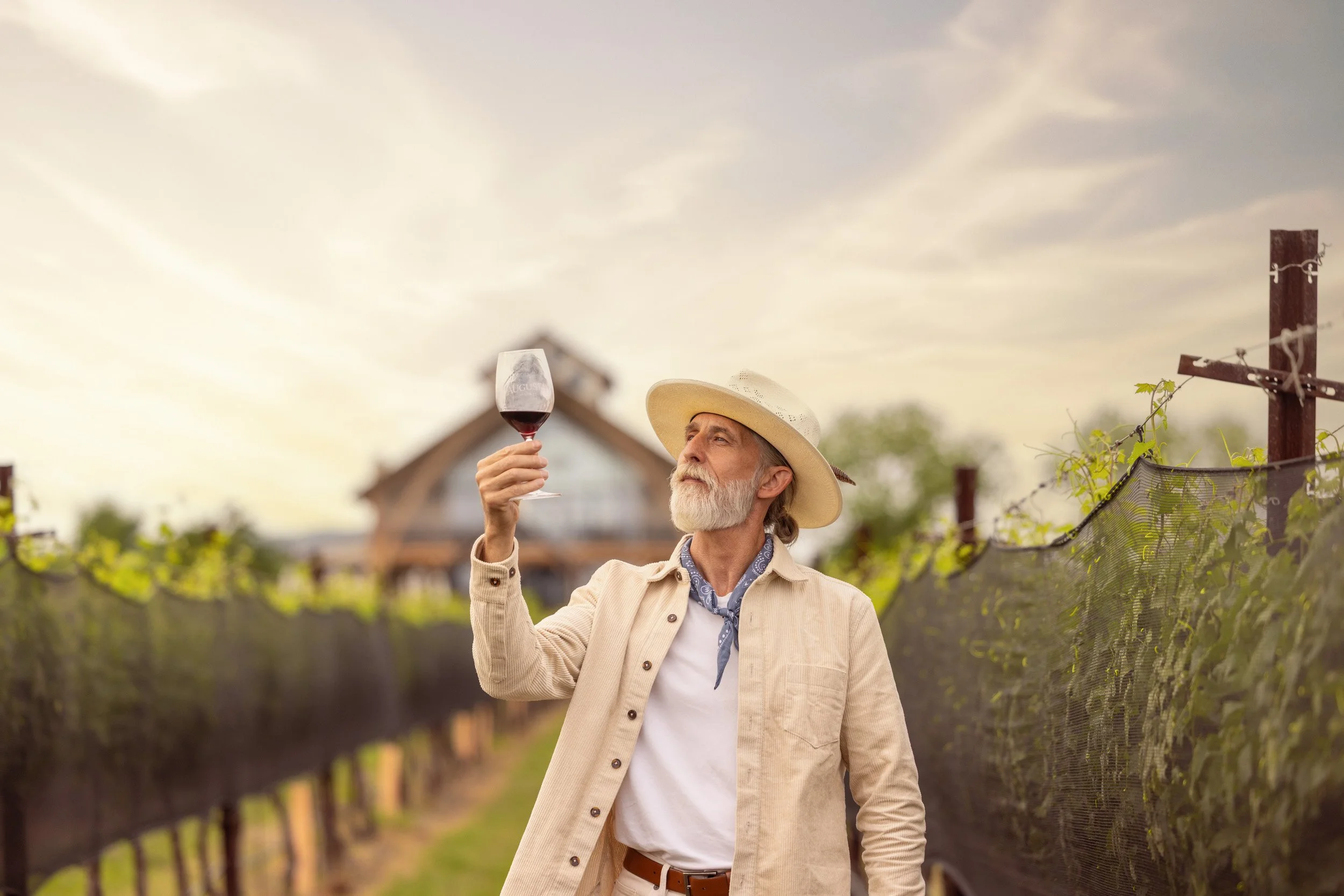 An older man with a gray beard and long hair, wearing a beige jacket, white shirt, patterned necktie, and a wide-brimmed hat, is holding a glass of red wine in an outdoor vineyard during sunset.