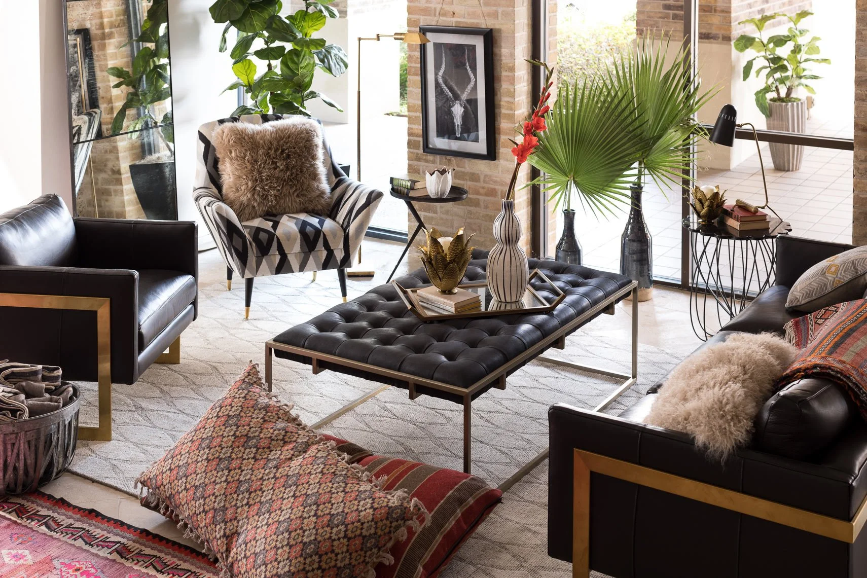 Modern living room with black leather sofas, an armchair with a fur pillow, assorted decorative pillows, a tufted black coffee table with vases and books, large green plants, framed artwork on brick wall, and a glass door leading outside.