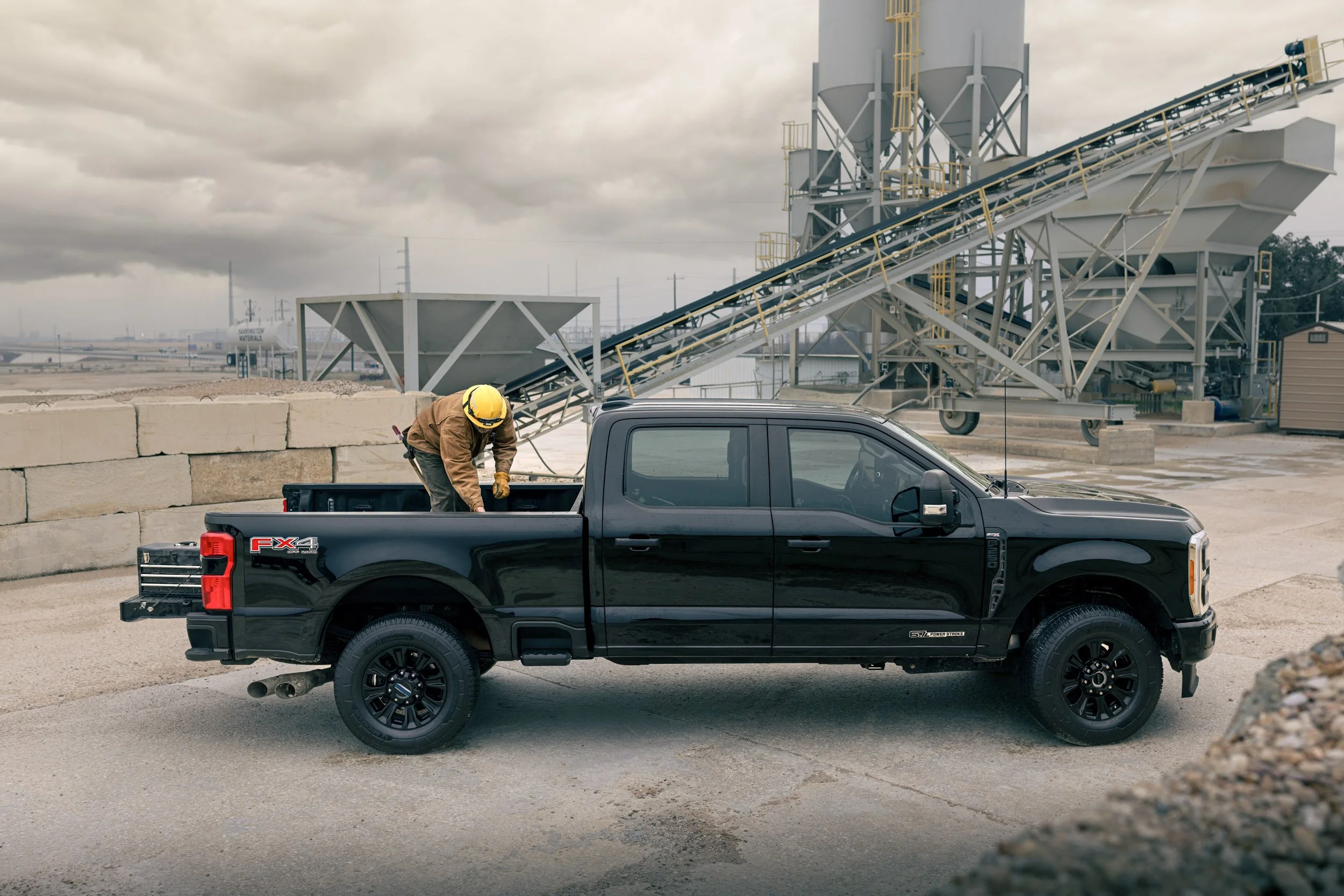 A man in a yellow safety helmet and brown jacket leaning into the bed of a black pickup truck, with large industrial structures in the background on a cloudy day.