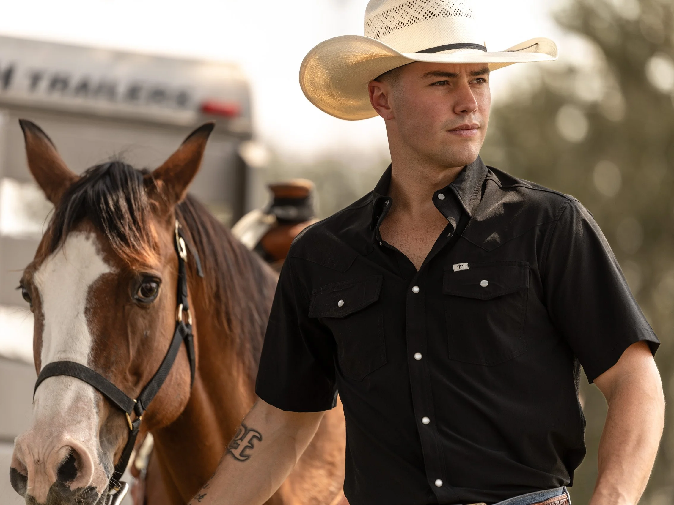 A young man wearing a cowboy hat and black shirt walking with a horse.