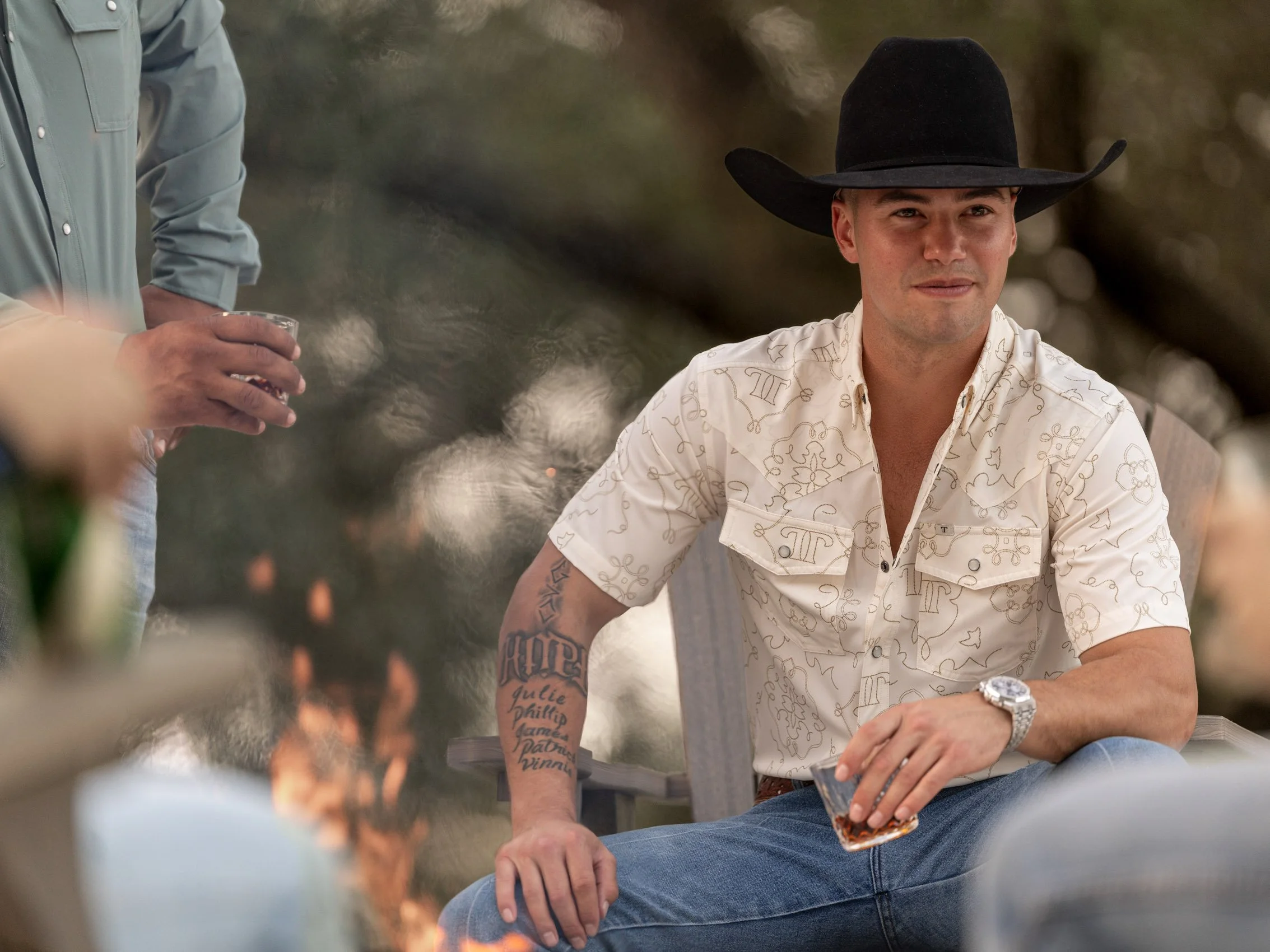 A young man wearing a black cowboy hat and a white shirt with a light pattern, sitting outdoors with a drink in hand, looking at the camera.