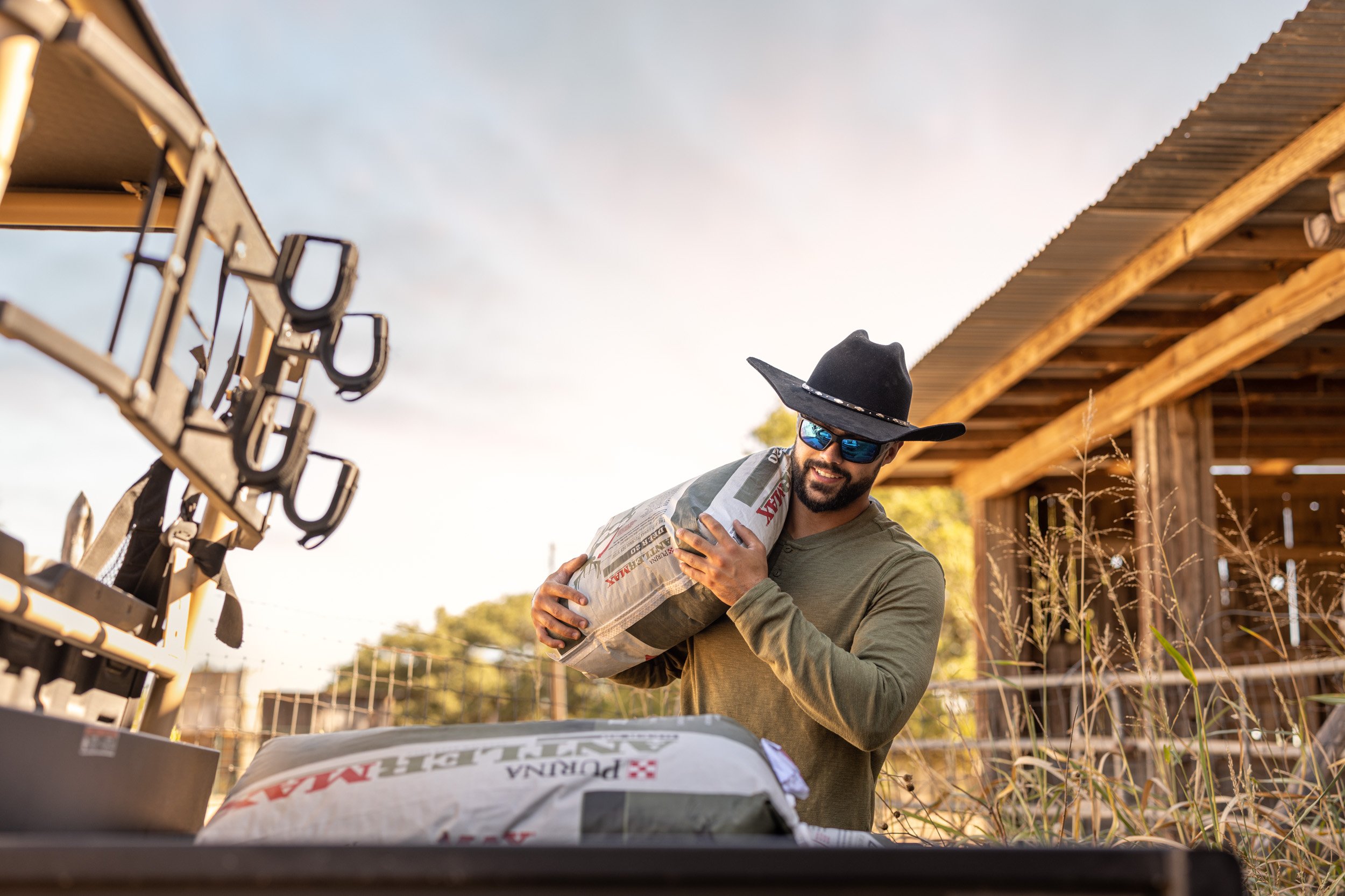 A man wearing sunglasses, a black cowboy hat, and a gray hoodie is holding a bag of fertilizer while smiling at a construction site or farm, with a wooden structure and tools or equipment nearby, during daytime.
