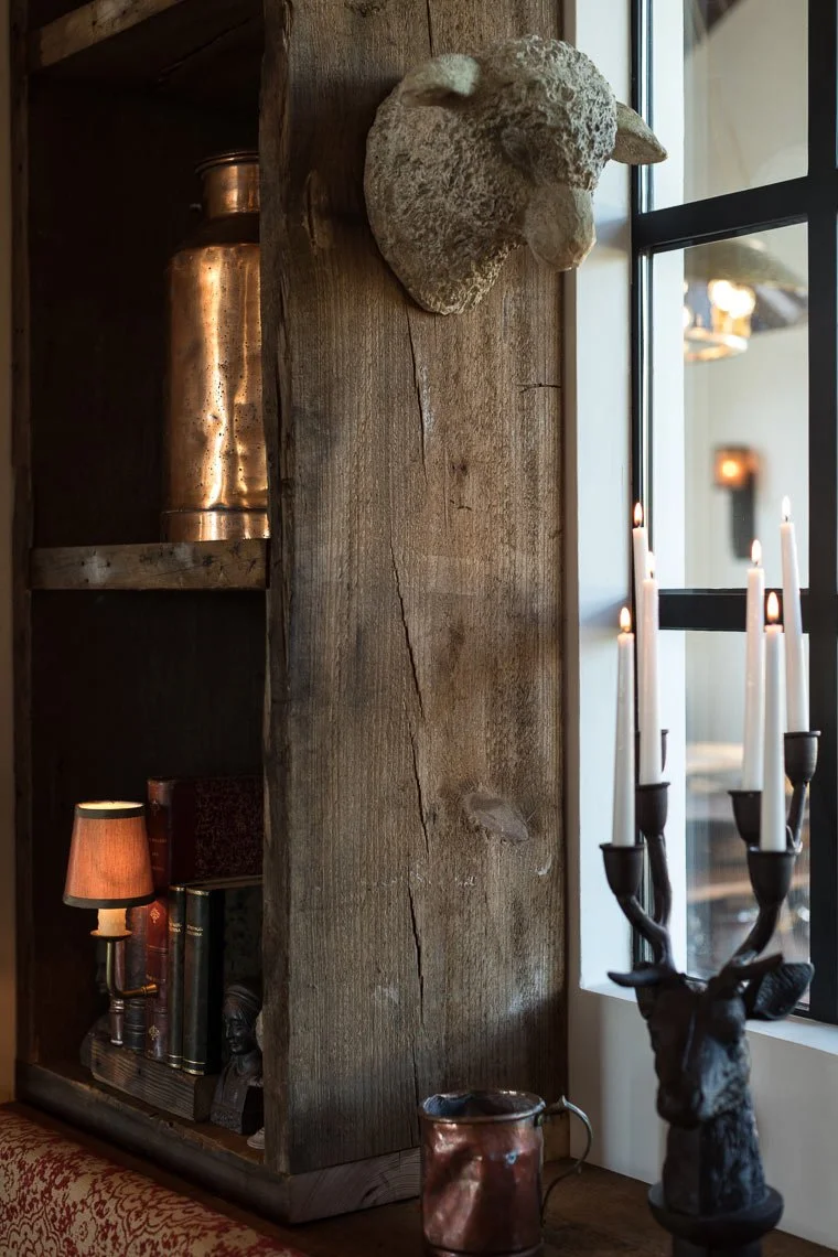 A decorative stone sheep head wall sculpture, a black metal candleholder with white candles, a copper mug, a small lamp, and a bookshelf with books and a small bust, all on a wooden surface next to a window.