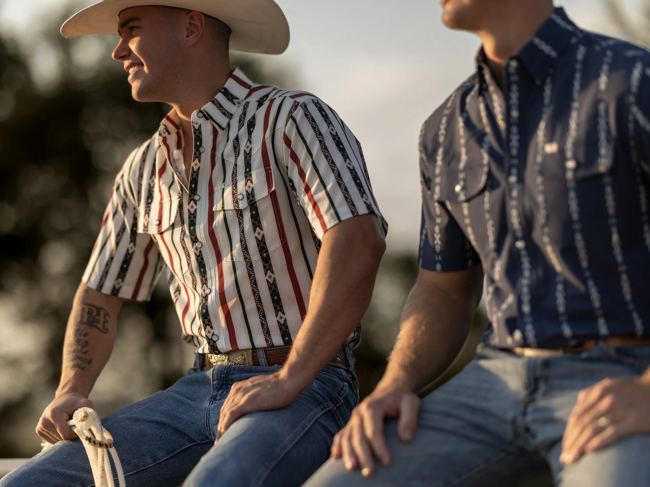 Two men sitting outdoors during sunset, wearing cowboy-style shirts with striped patterns, jeans, and cowboy hats, engaging in conversation.