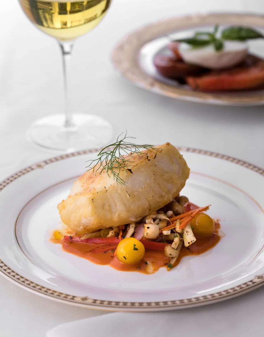 A white plate with a baked fish fillet garnished with a sprig of dill, served over a bed of mixed vegetables including cherry tomatoes and shredded carrots, with a brown sauce. In the background, there is a glass of white wine and a plate with additi