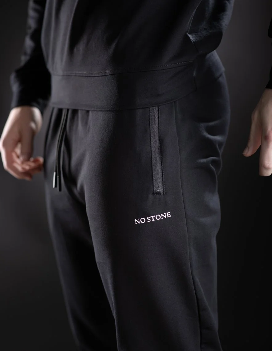 Close-up of a person wearing black athletic pants with the text 'NO STONE' on the thigh, and a black zippered pocket.