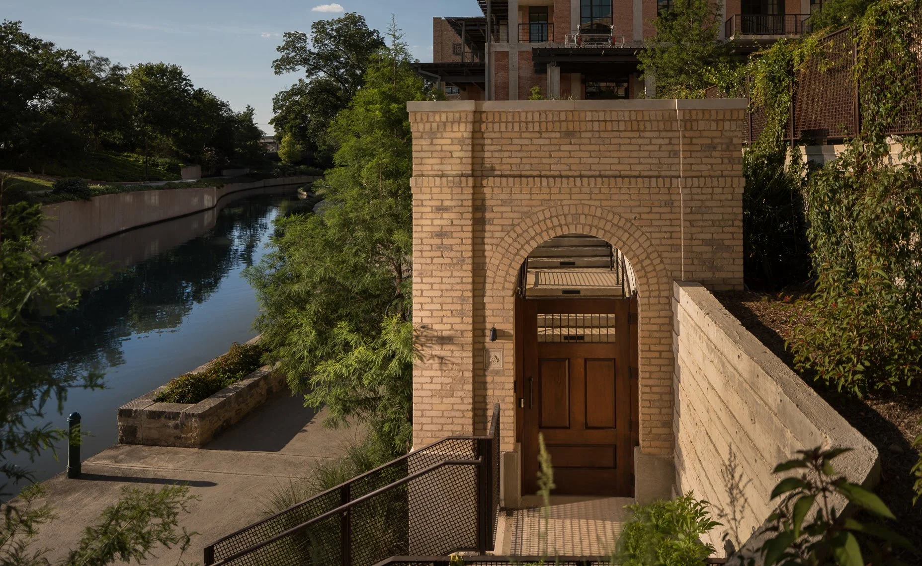 Brick archway with a wooden gate in front of a sidewalk, overlooking a canal with lush trees on the opposite bank, and modern buildings under a clear sky.