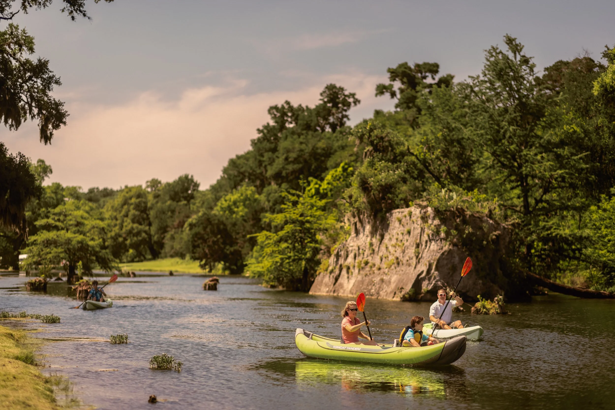 People kayaking on a river with lush green trees and rocky cliffs in the background on a sunny day.