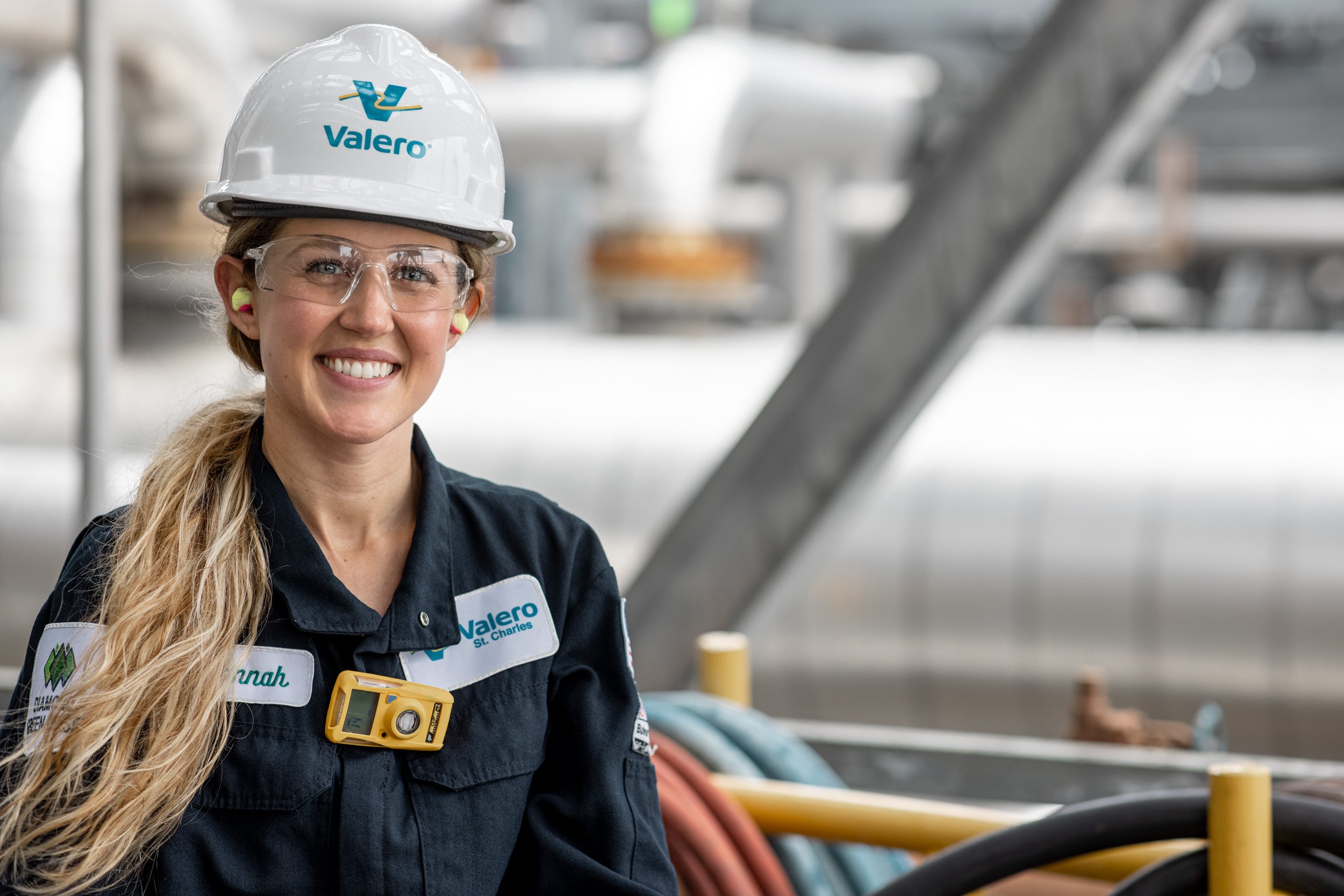 A woman wearing a white safety helmet with the Valero logo, clear safety glasses, and a navy uniform with Valero patches smiling at the camera inside an industrial facility with colorful pipes and safety barriers.