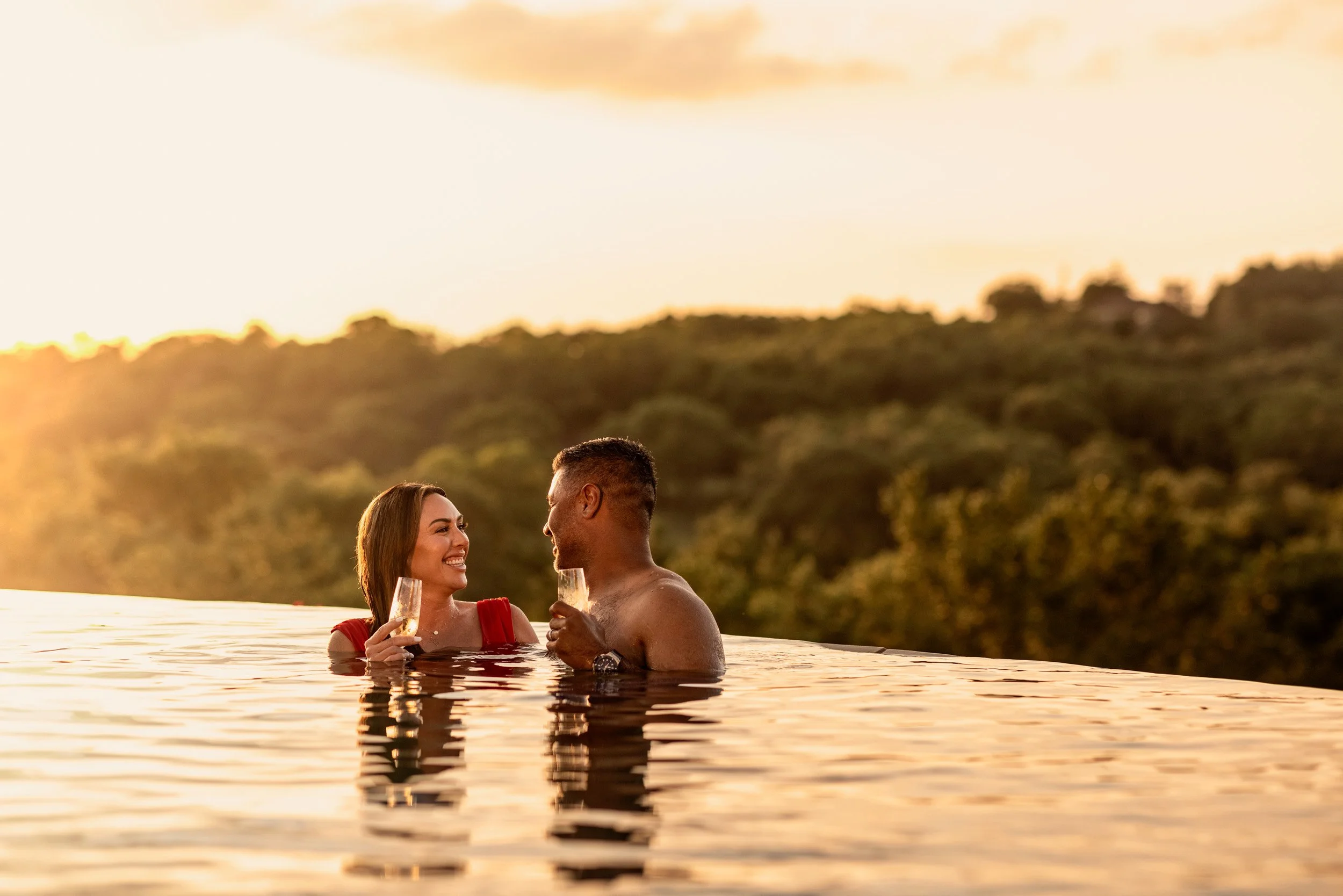 A couple enjoying drinks and smiling while sitting in an outdoor hot tub or pool during sunset, with a scenic wooded landscape in the background.